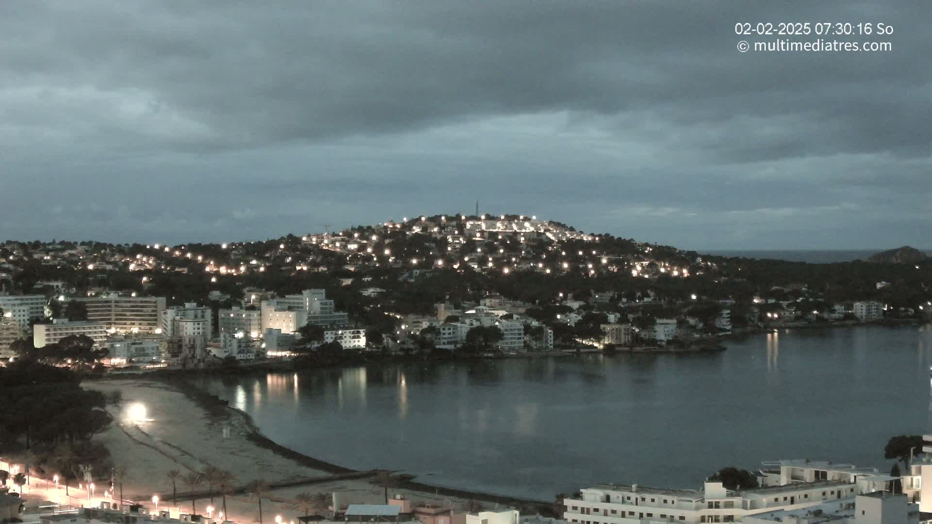 A nighttime view of a coastal town illuminated by lights, nestled around a calm bay under a cloudy sky.
