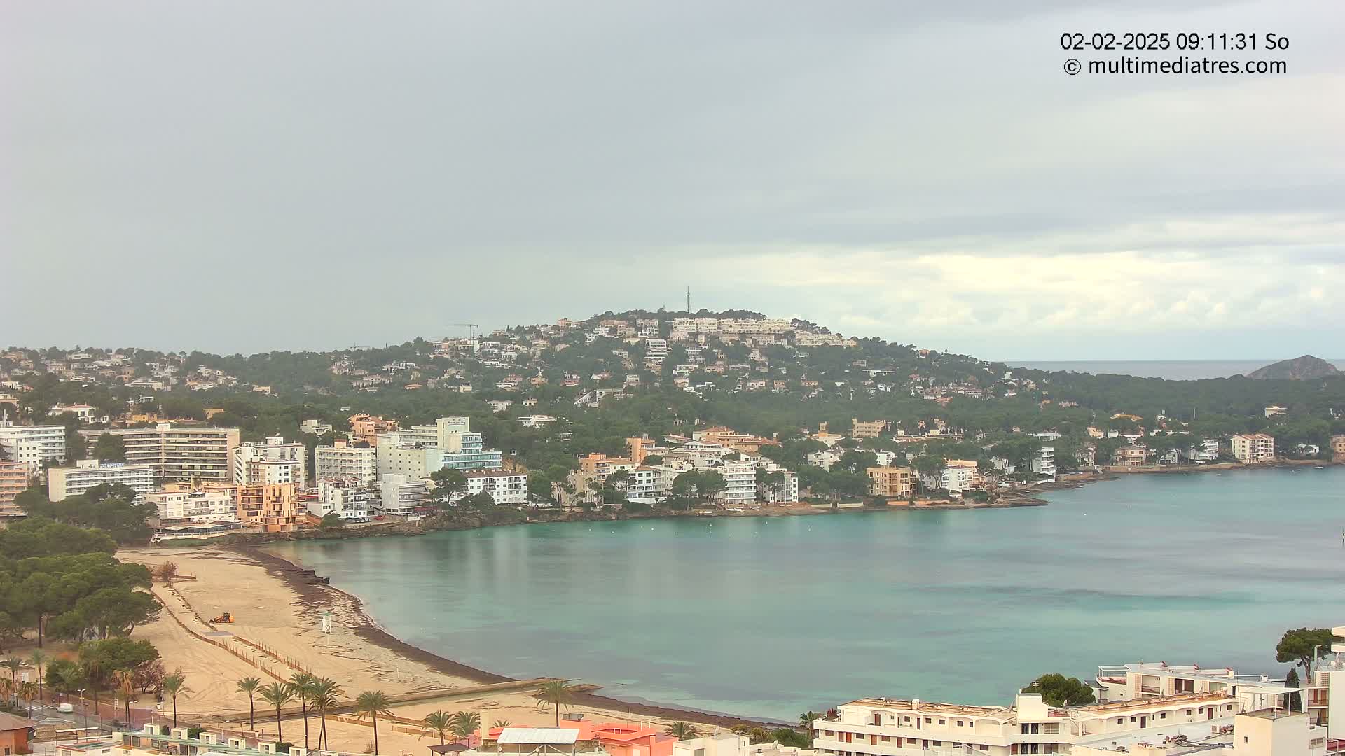 A coastal town with multi-story buildings sits on a hill overlooking a calm bay under an overcast sky.