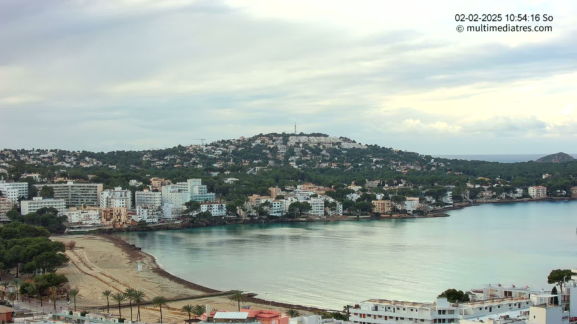 A coastal town with a sandy beach and numerous buildings sits nestled against a green hill under a cloudy sky.