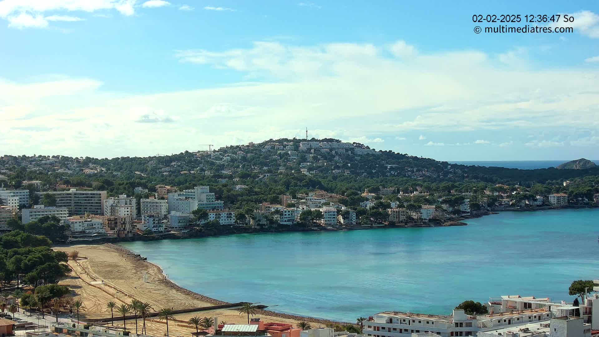 A coastal town nestled against a hill, featuring a sandy beach and calm, turquoise waters under a partly cloudy sky.