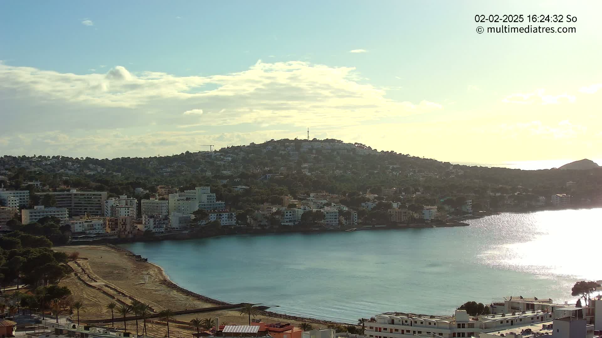 A coastal town nestled against a hill under a partly cloudy sky, with a calm body of water reflecting sunlight.