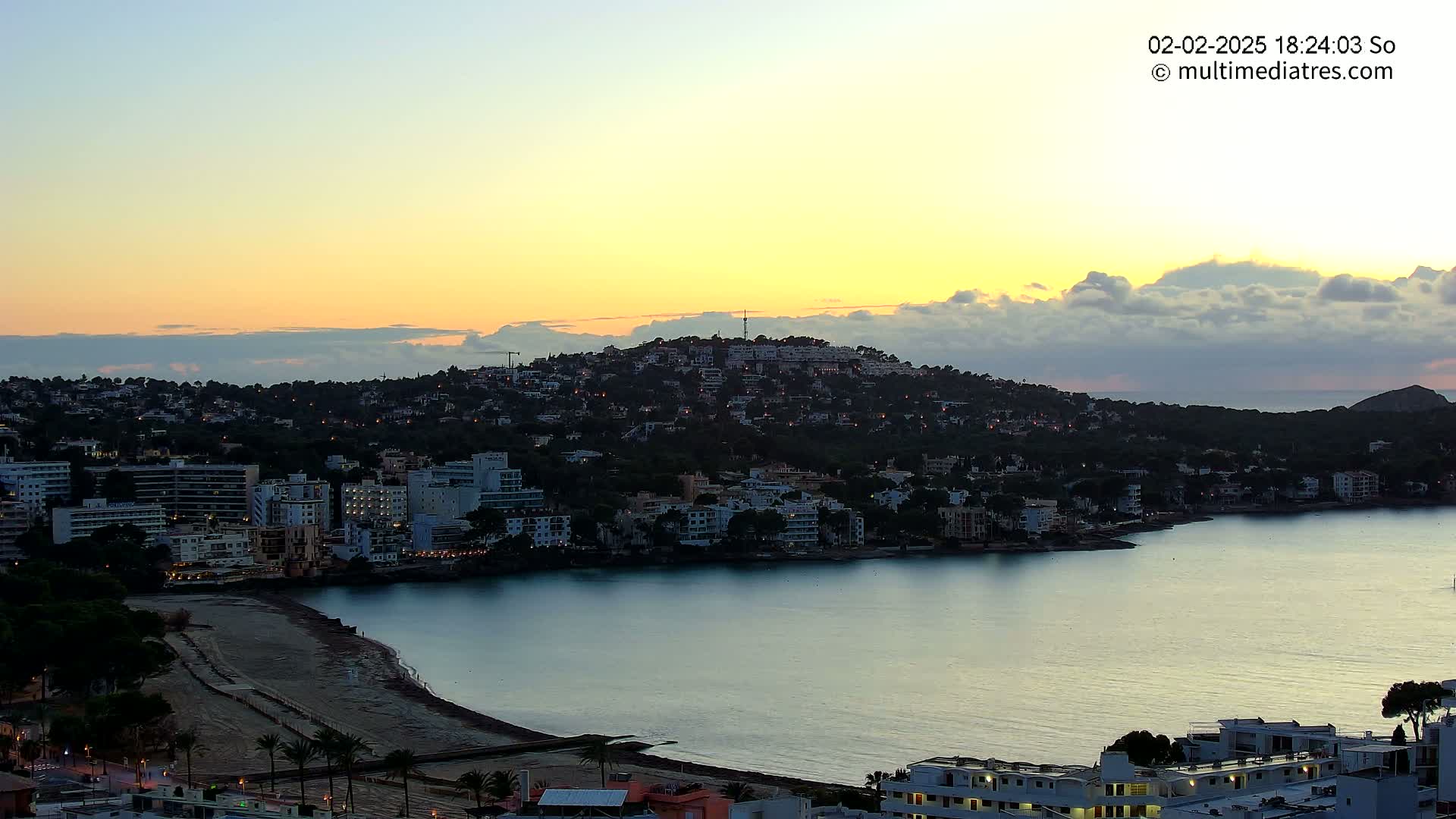 A coastal town nestled against a hill at sunset, with a calm bay stretching out to the horizon.