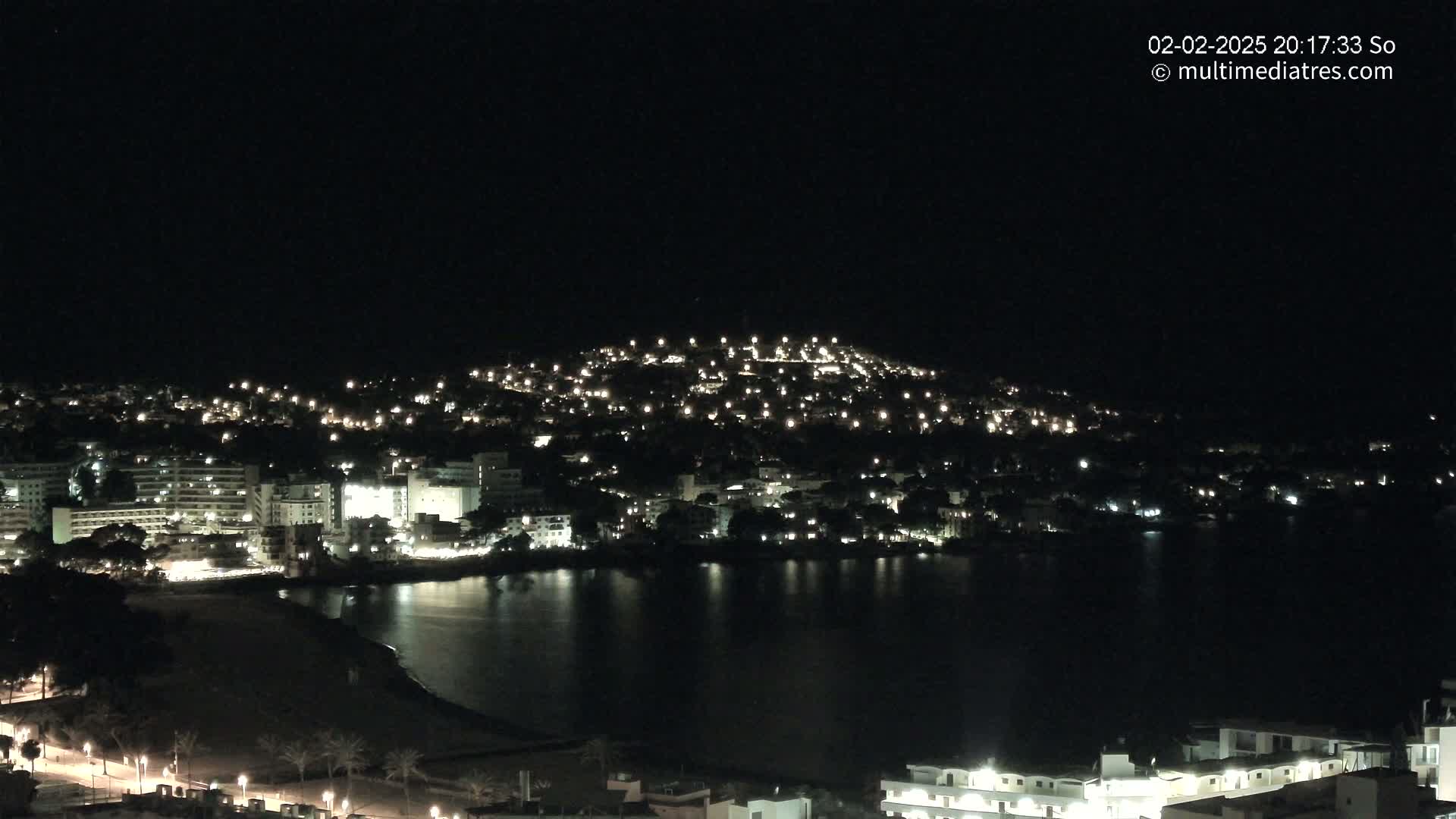 A nighttime view of a coastal town with numerous lights illuminating buildings and streets, beside a dark body of water under a clear, dark sky.