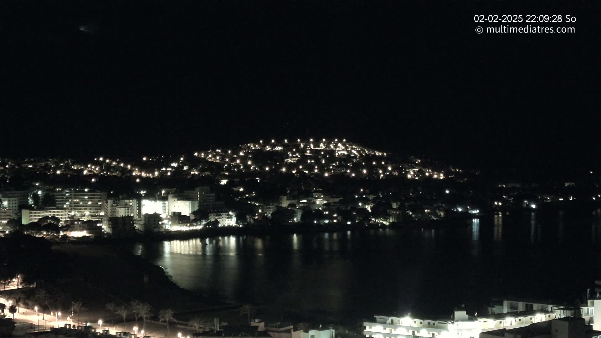 A nighttime view of a coastal town illuminated by lights, reflected in a calm body of water under a clear, dark sky.