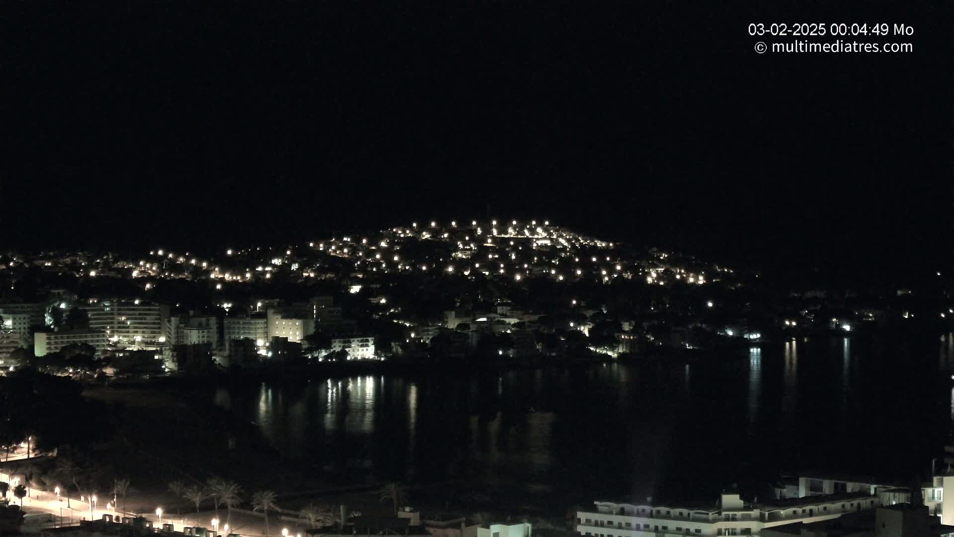A nighttime view of a coastal town illuminated by numerous lights, reflecting on a dark body of water under a clear sky.