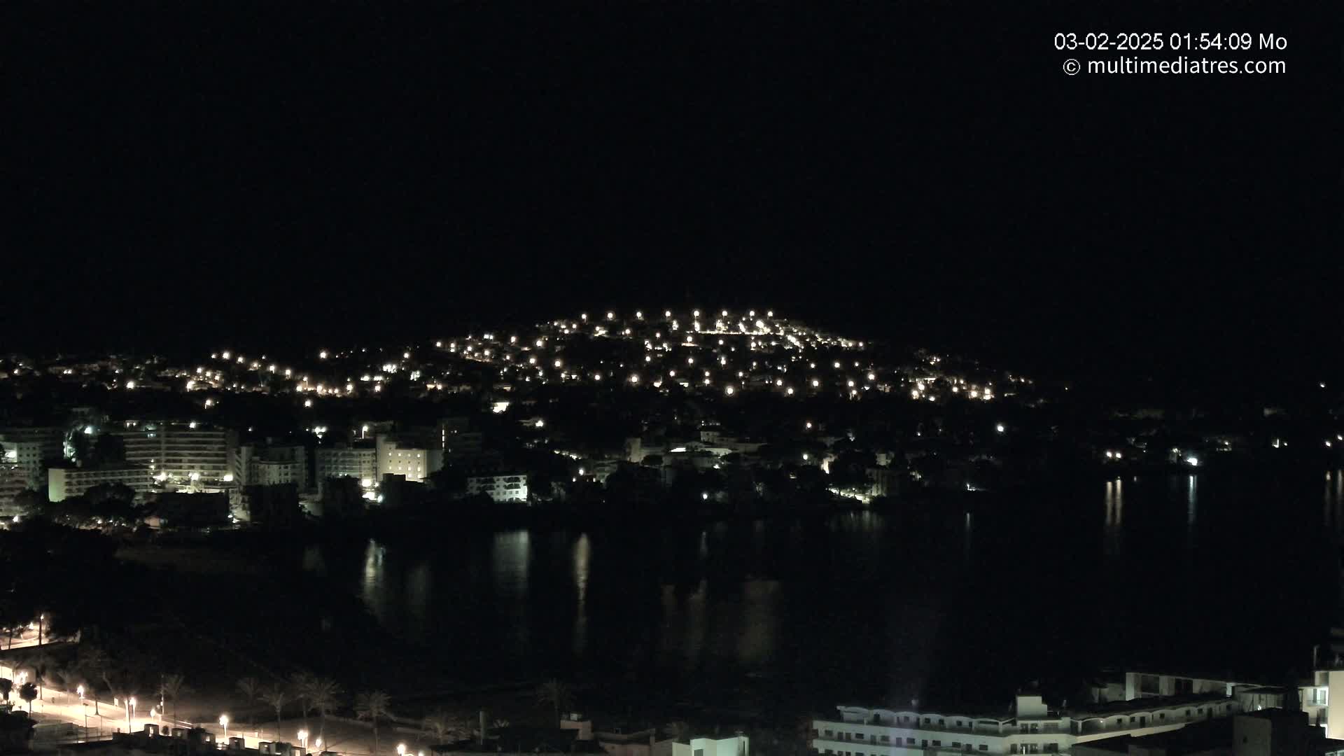 A nighttime view of a city illuminated by scattered lights, situated beside a dark body of water under a clear, dark sky.