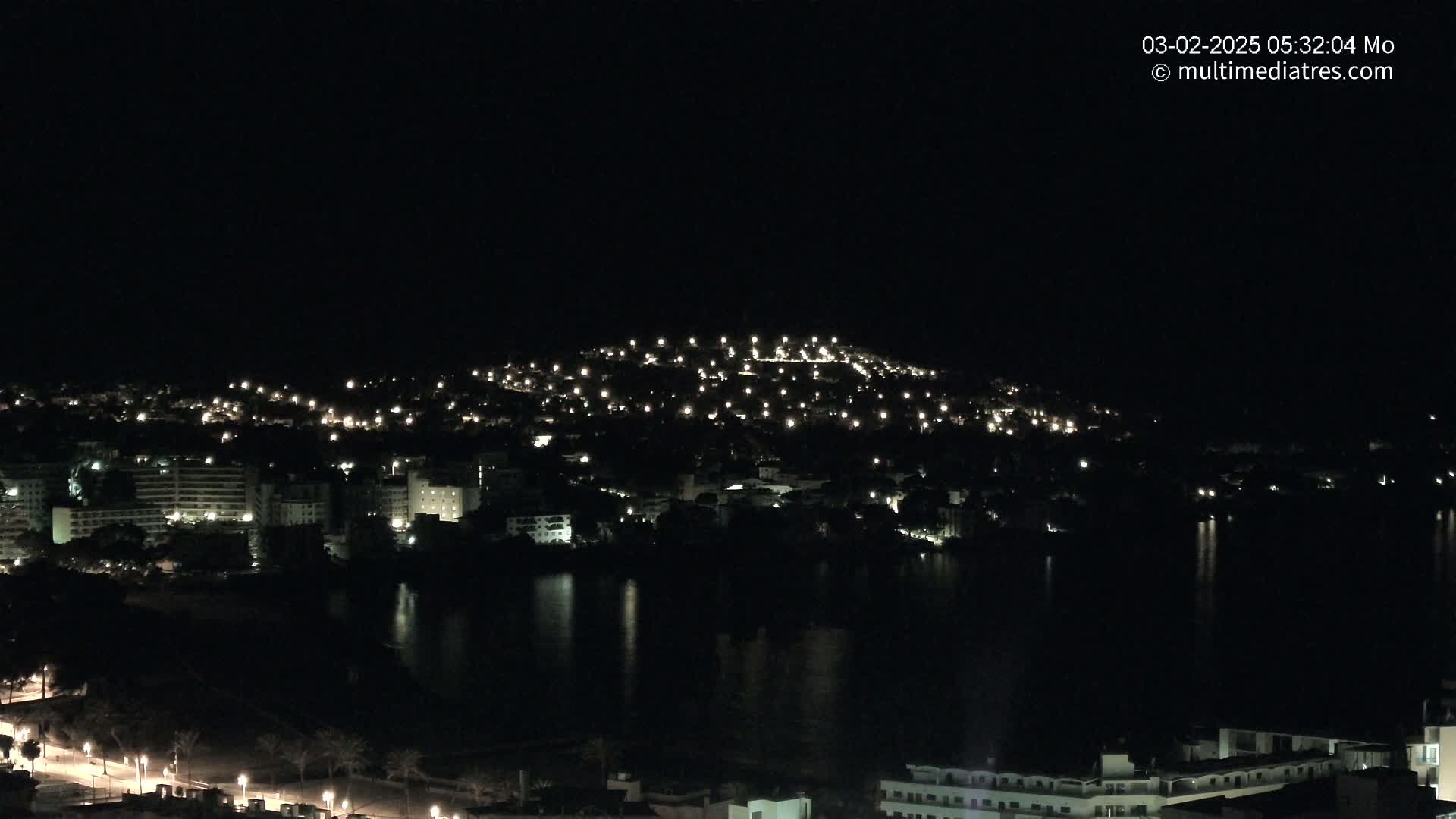 A nighttime view of a coastal town illuminated by numerous lights, reflected in a dark body of water under a clear, dark sky.