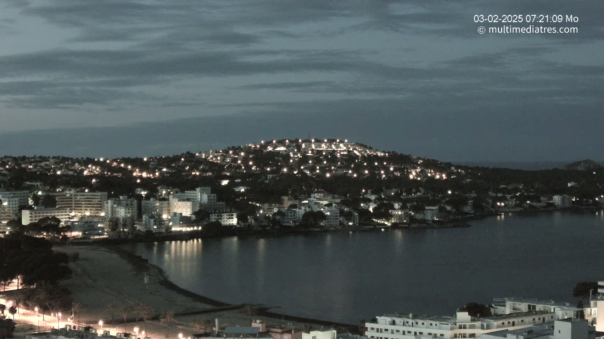 A nighttime view of a coastal town illuminated by lights, nestled against a dark hill, overlooking a calm bay under a cloudy sky.