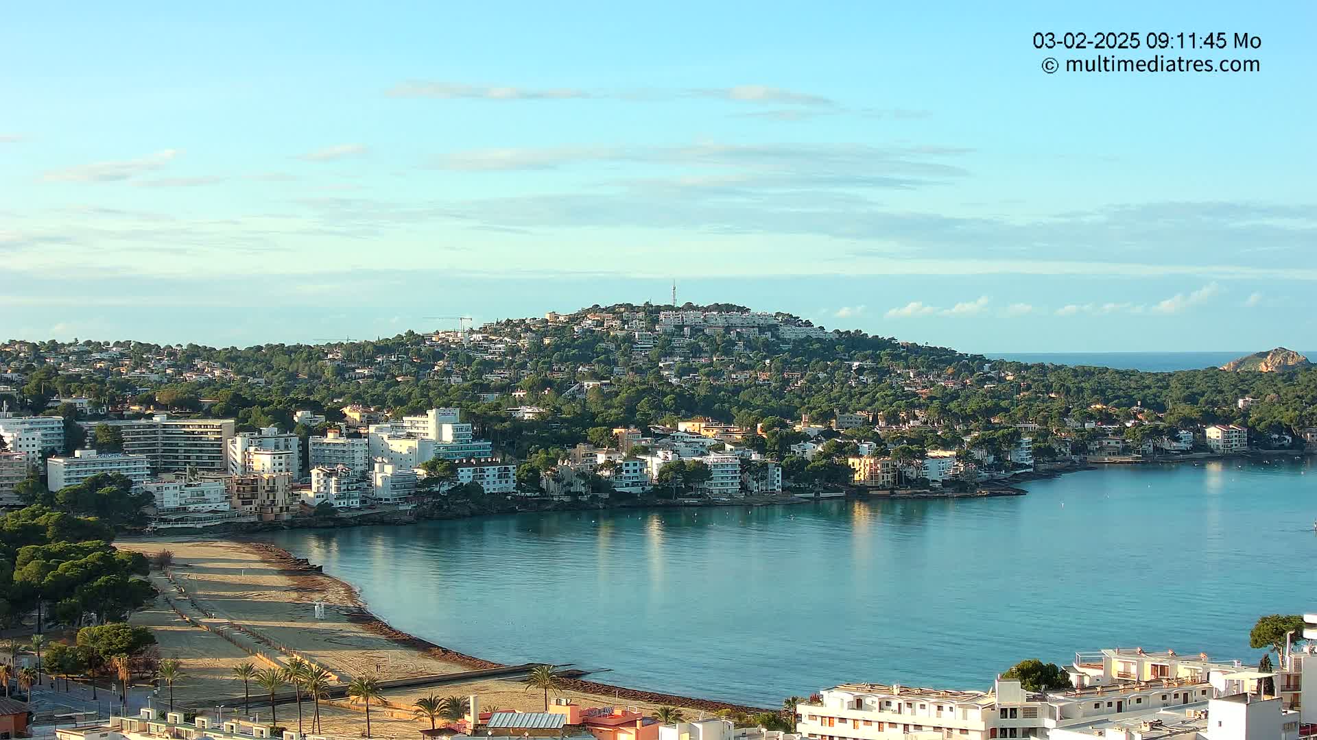 A coastal town with a sandy beach and calm, clear water sits nestled below a hill covered in buildings and green trees under a mostly clear sky.