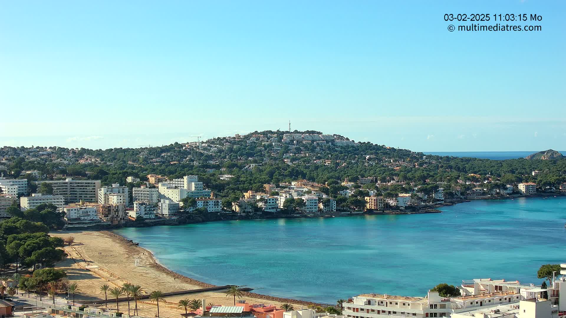 A coastal town with turquoise water, a sandy beach, and buildings nestled on a hillside under a clear blue sky.