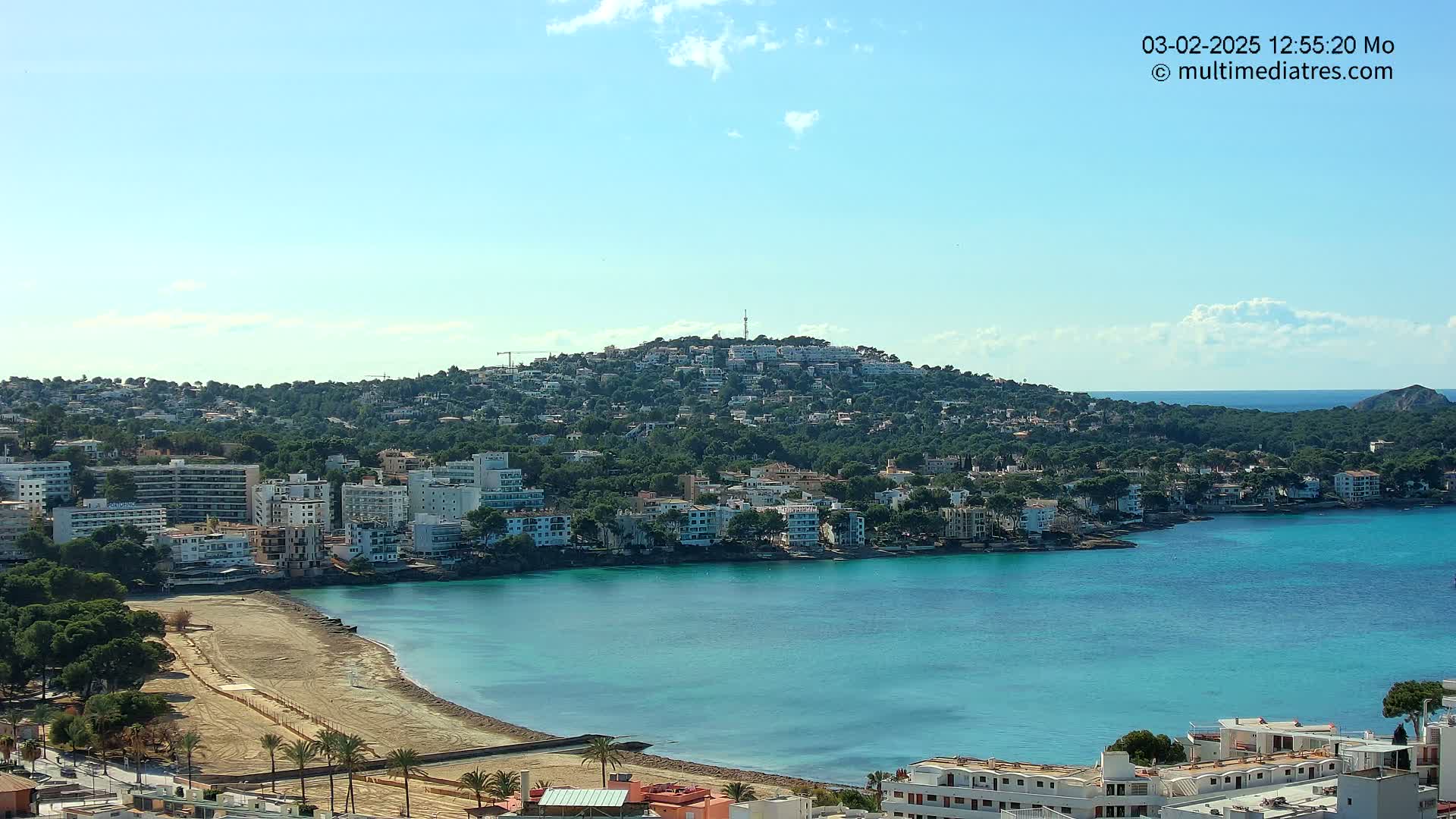 A clear, sunny day reveals a coastal town nestled against a verdant hill, with a sandy beach and calm, turquoise water forming a tranquil bay.