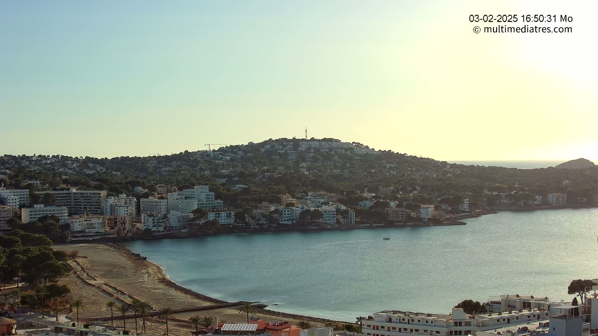 A coastal town nestled against a hill overlooking a calm, blue bay under a clear, light sky.