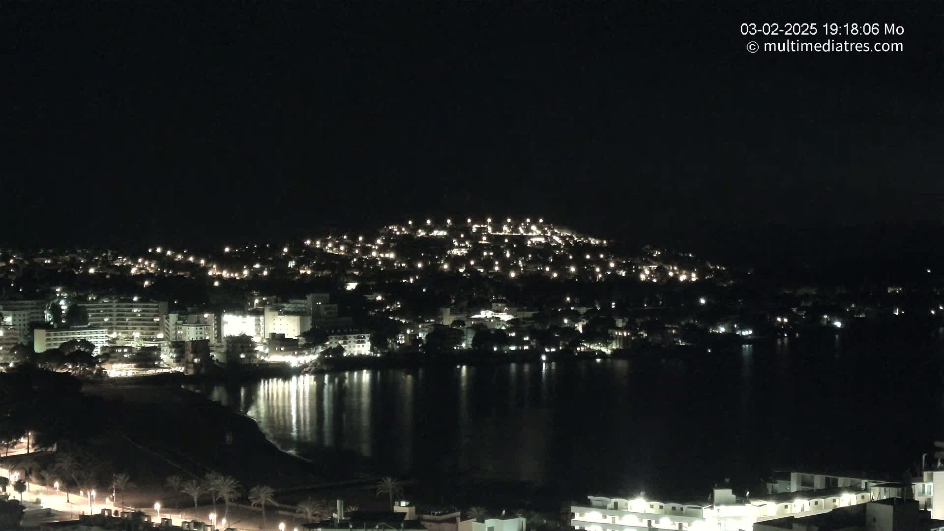 A nighttime view of a coastal town illuminated by numerous lights, reflecting on a dark body of water under a clear sky.