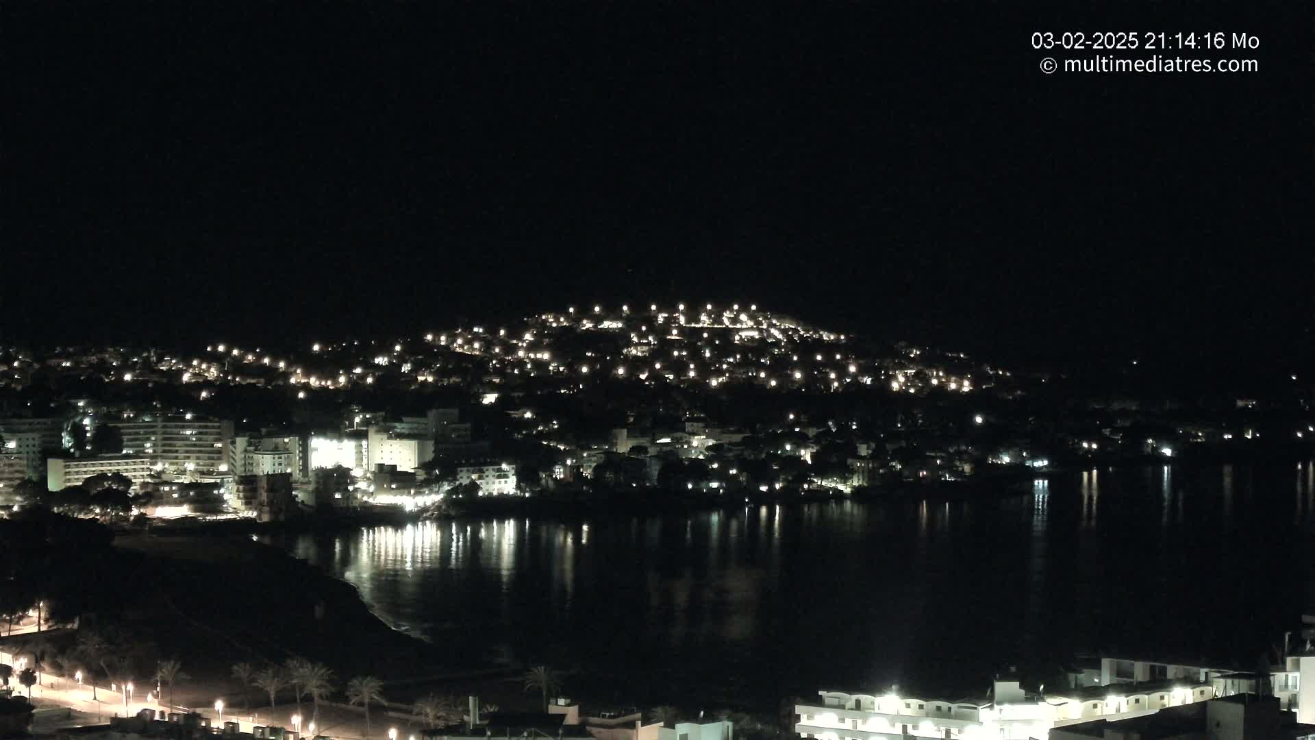A nighttime view of a city situated on a hillside overlooking a calm body of water, with the city lights reflecting on the water's surface.