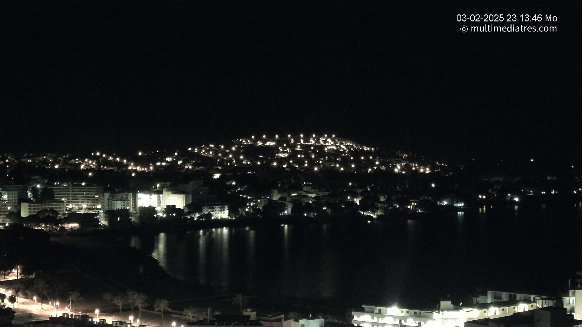 A nighttime view of a town illuminated by lights, situated beside a dark body of water, under a clear sky.