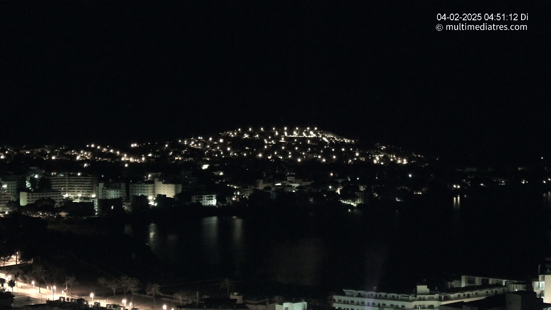 A nighttime view of a city illuminated by scattered lights, situated near a dark body of water under a clear, dark sky.