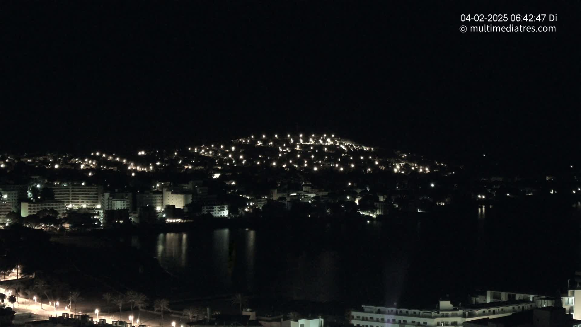 A nighttime view of a coastal town with numerous lights illuminating buildings and streets, reflected in a dark body of water.