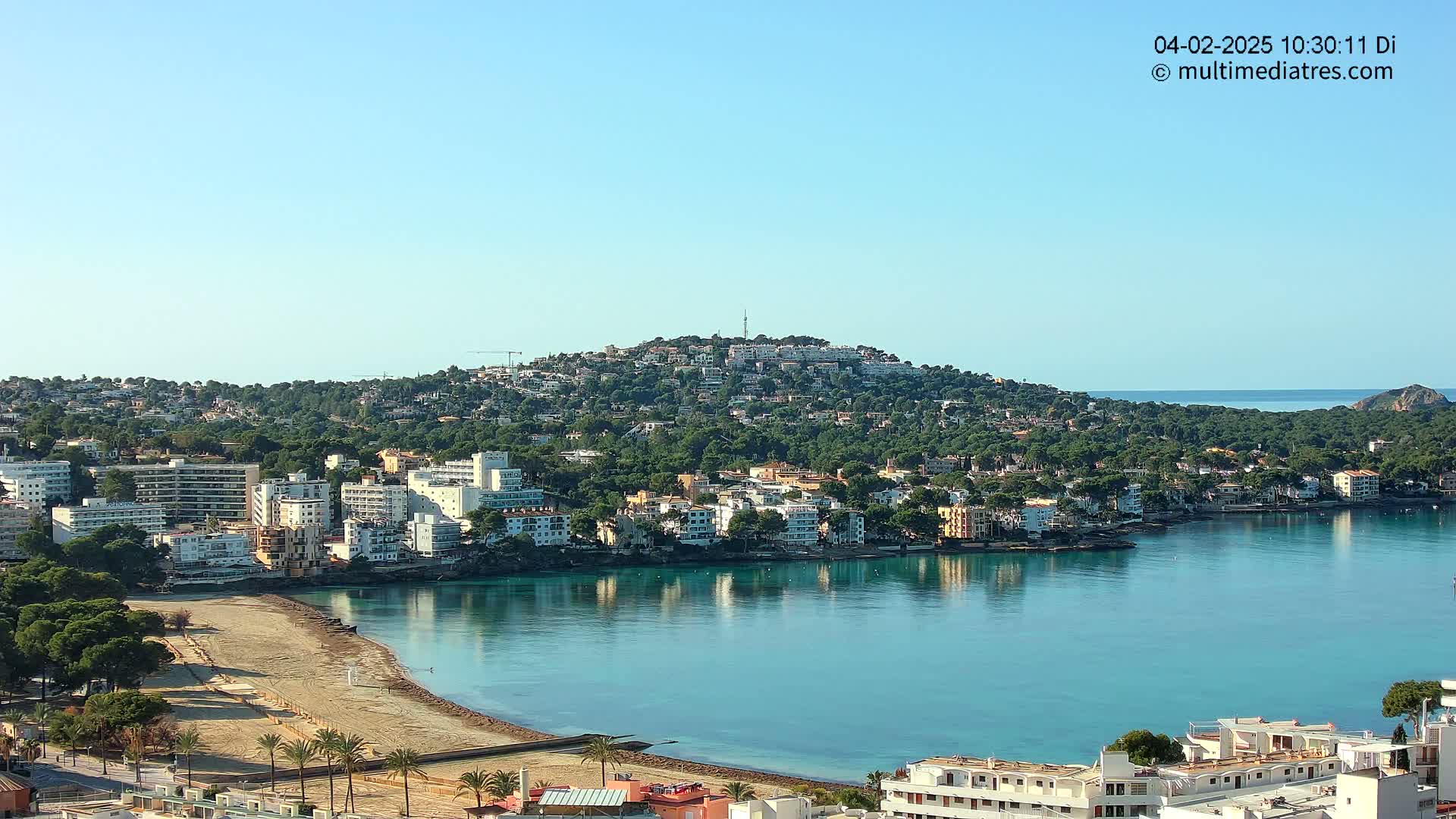 A coastal town nestled beside a calm, turquoise bay, backed by a hill covered in Mediterranean vegetation under a clear blue sky.