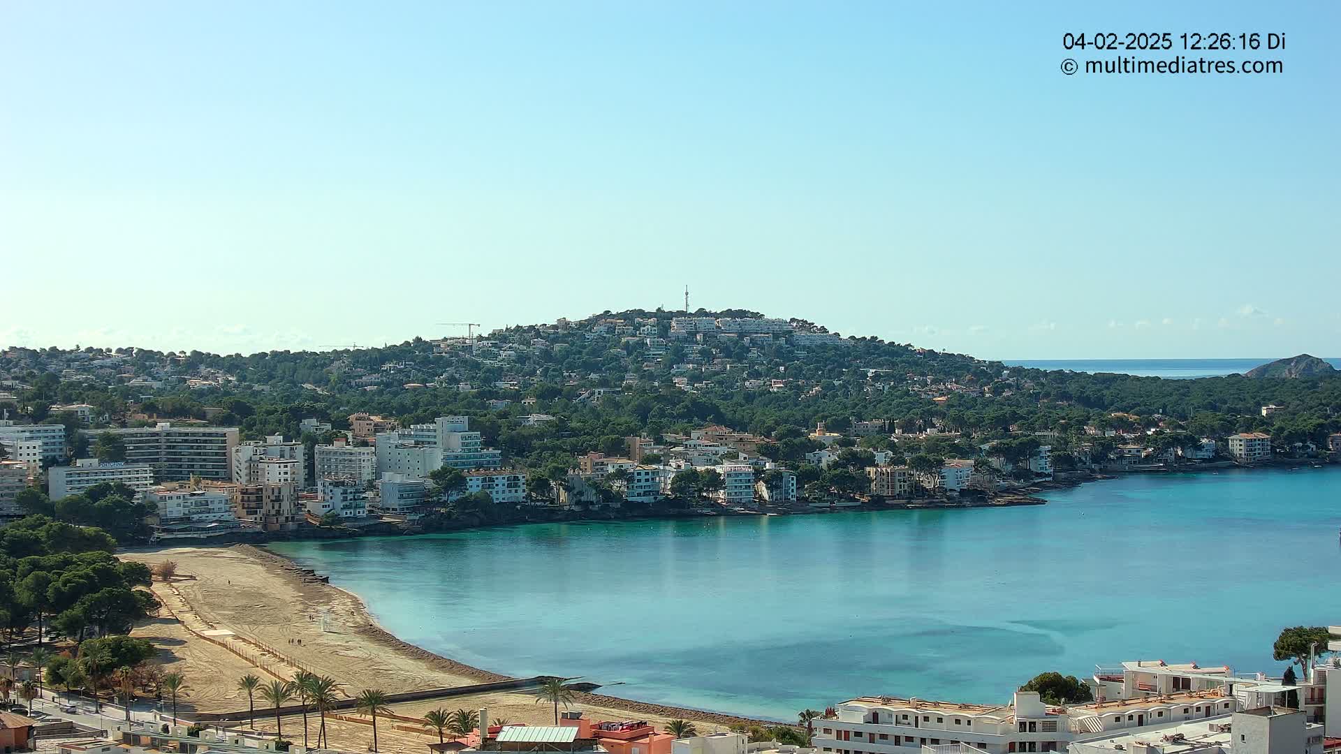 A coastal town with a sandy beach and turquoise water, nestled against a hill covered in buildings and trees under a clear blue sky.