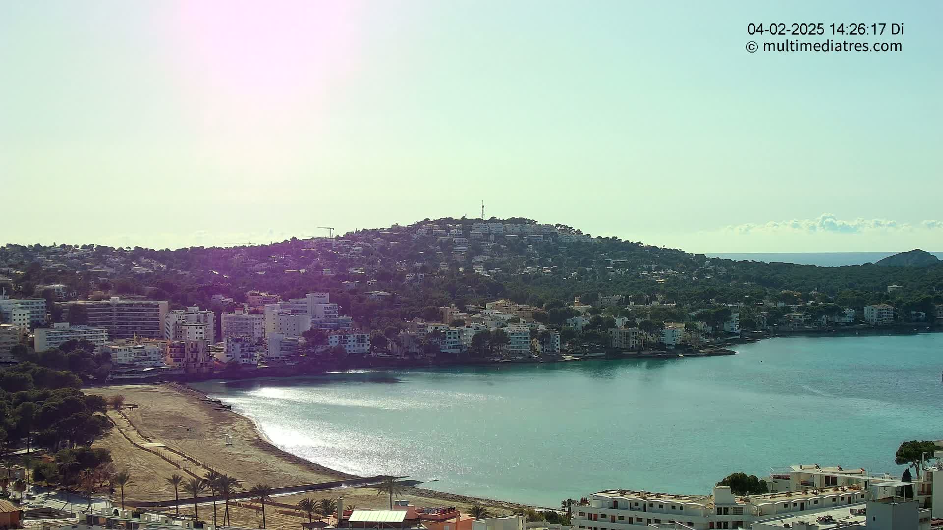 A coastal town nestled against a hill, with a calm, turquoise bay under a mostly clear sky.