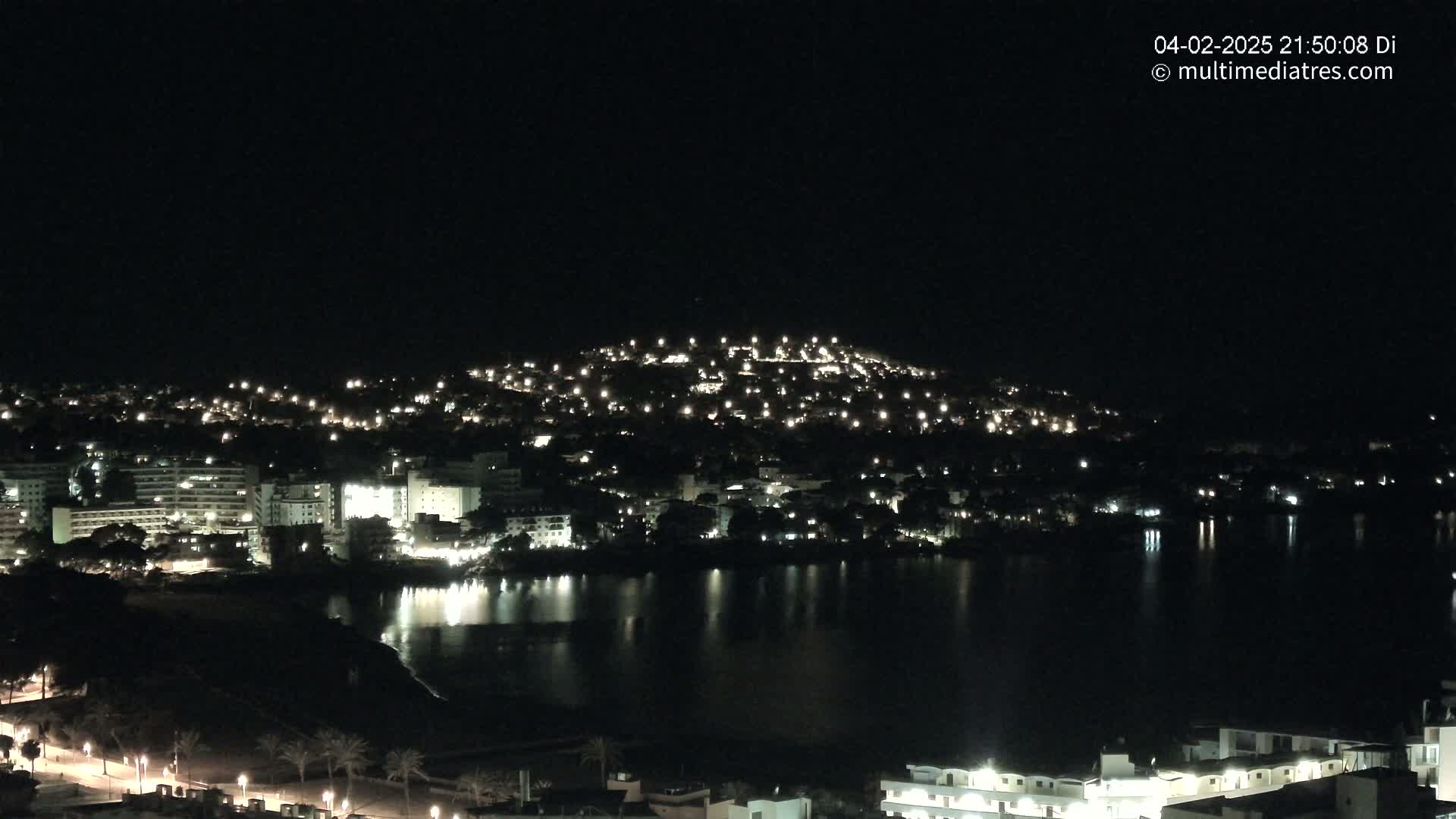 A nighttime view of a city illuminated by many lights nestled beside a dark body of water under a clear, dark sky.