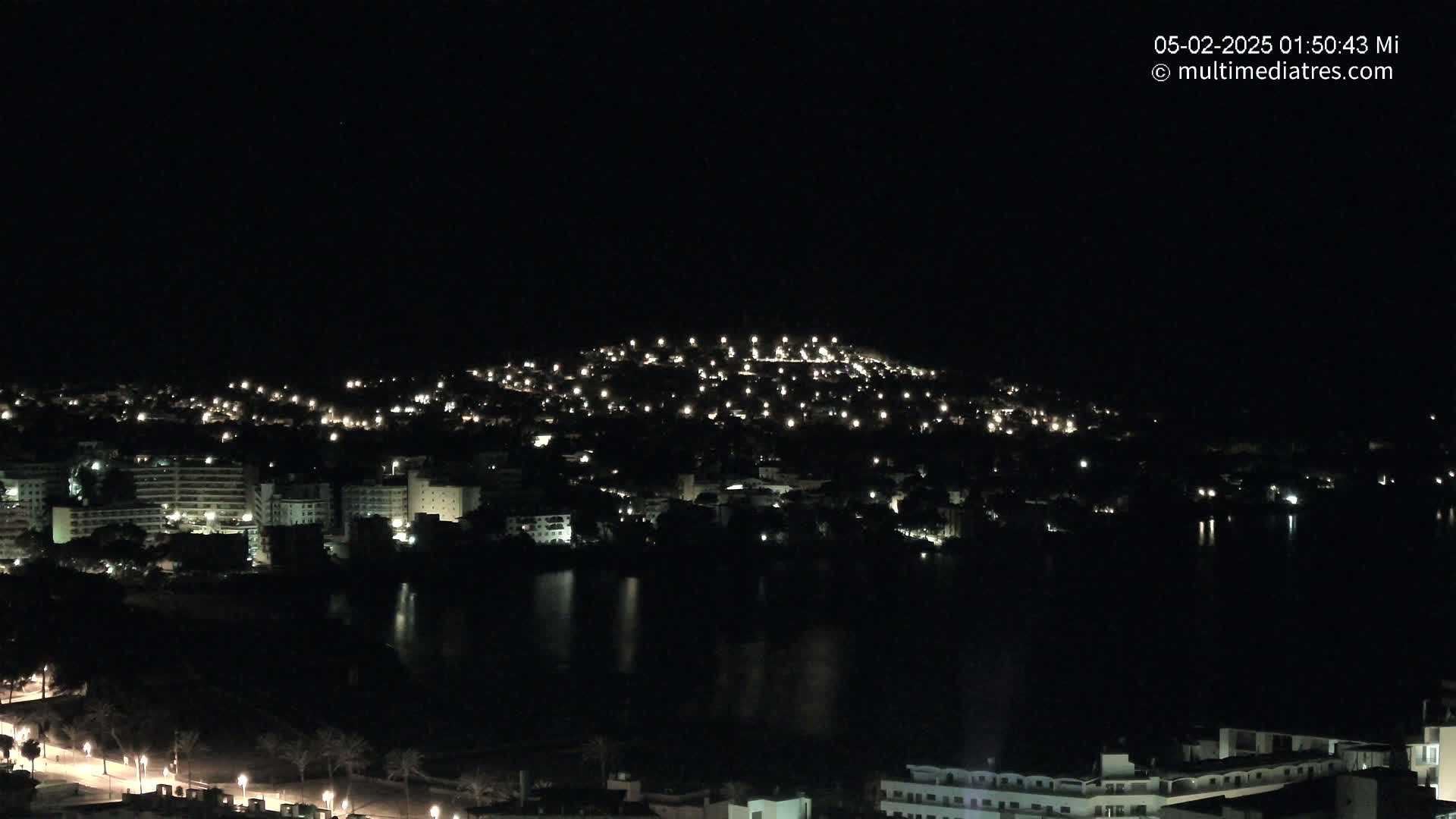 A nighttime view of a coastal town illuminated by numerous lights, with a dark body of water in the foreground.