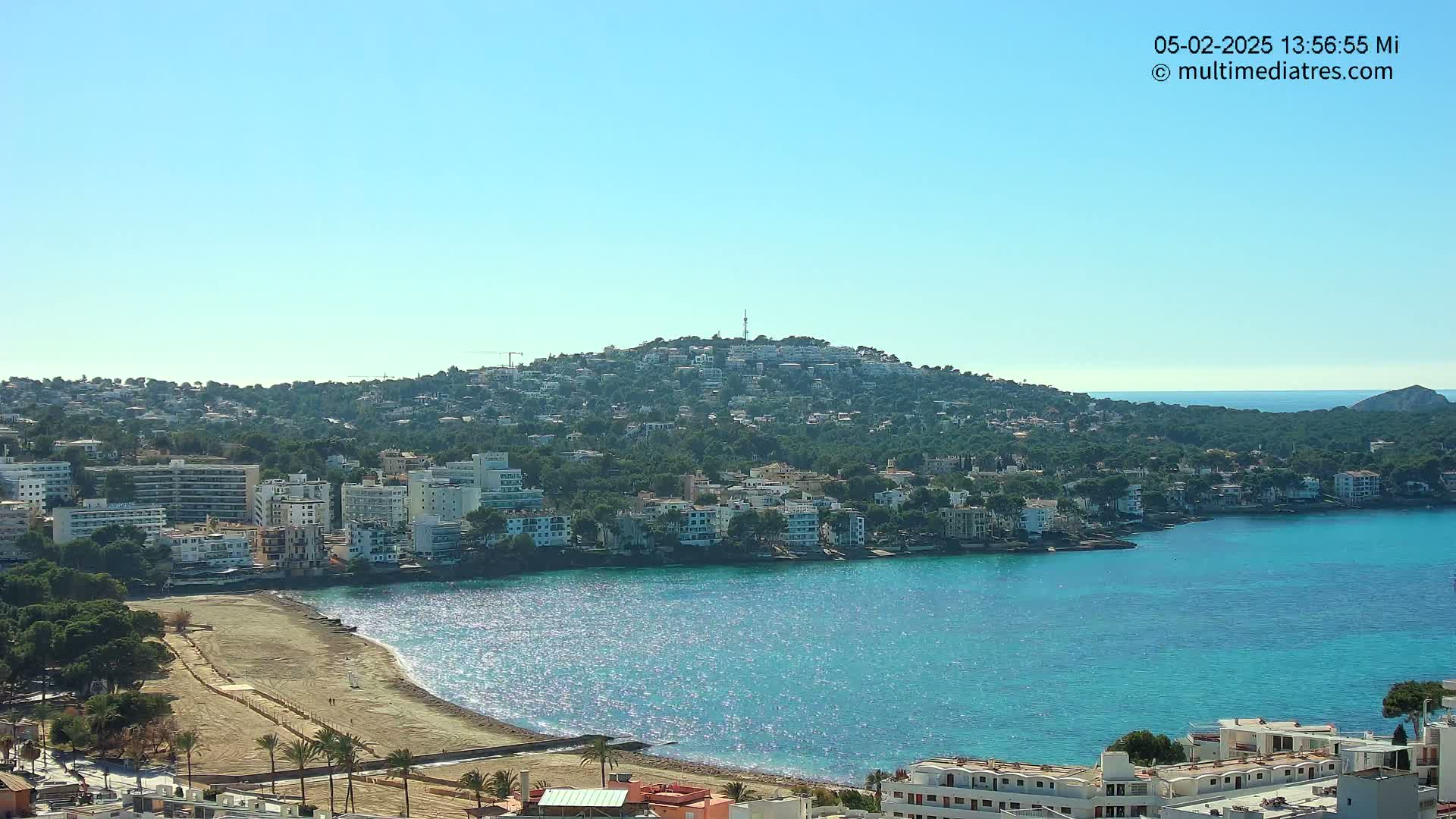 A hilltop town overlooks a calm, sparkling blue bay with a sandy beach, under a clear blue sky.