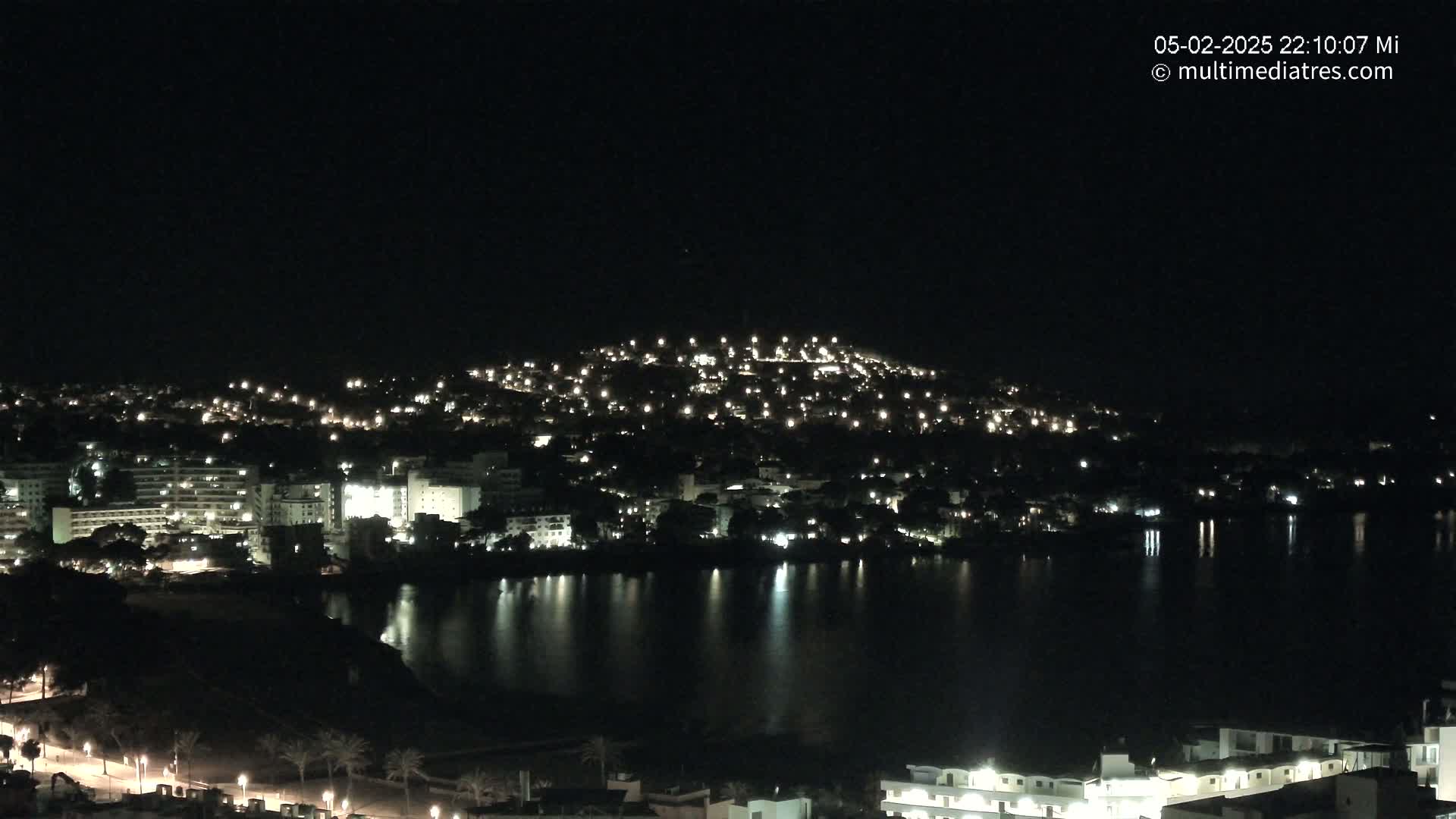 A nighttime view of a coastal town illuminated by lights, reflecting on a calm body of water.