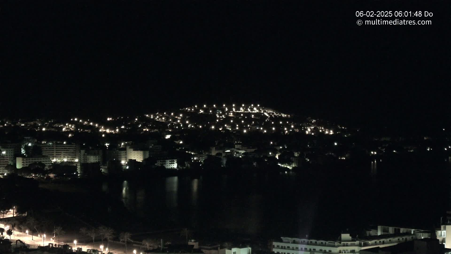 A nighttime view of a coastal town, illuminated by numerous lights, with a dark body of water reflecting the lights.