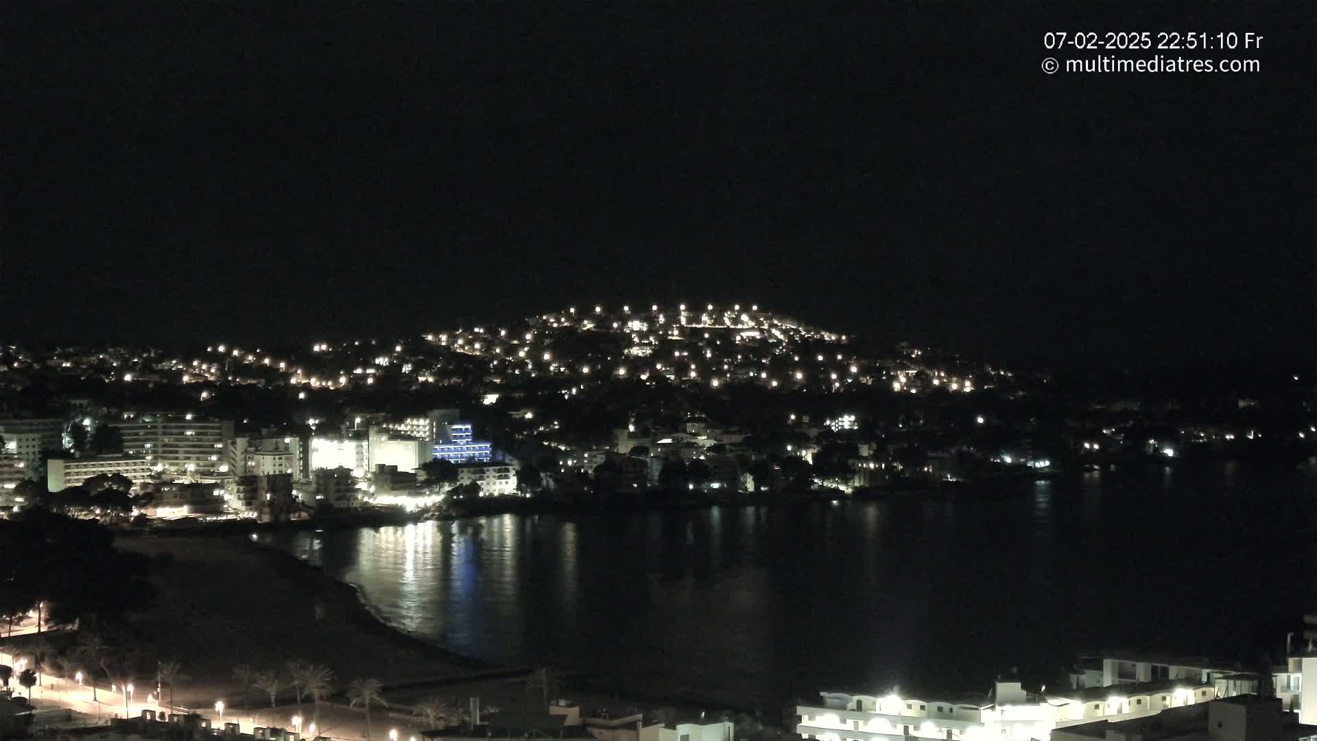 A nighttime view of a coastal town illuminated by lights, with calm water reflecting the city lights.