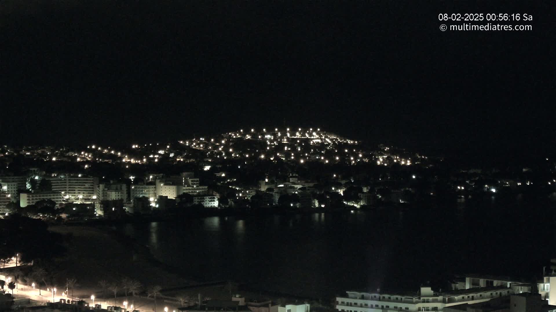 A nighttime view of a coastal town with numerous lights illuminating buildings and streets, nestled beside a dark body of water under a clear, dark sky.