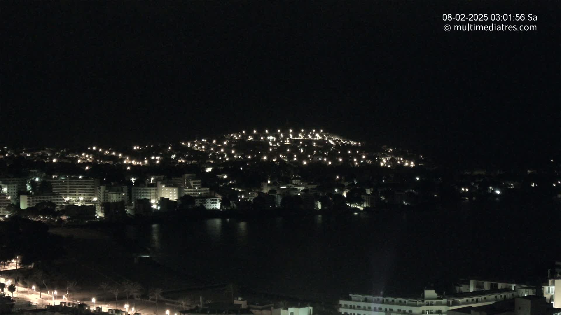 A nighttime view of a city spread across a hill and along a dark body of water, with numerous lights illuminating buildings and streets.