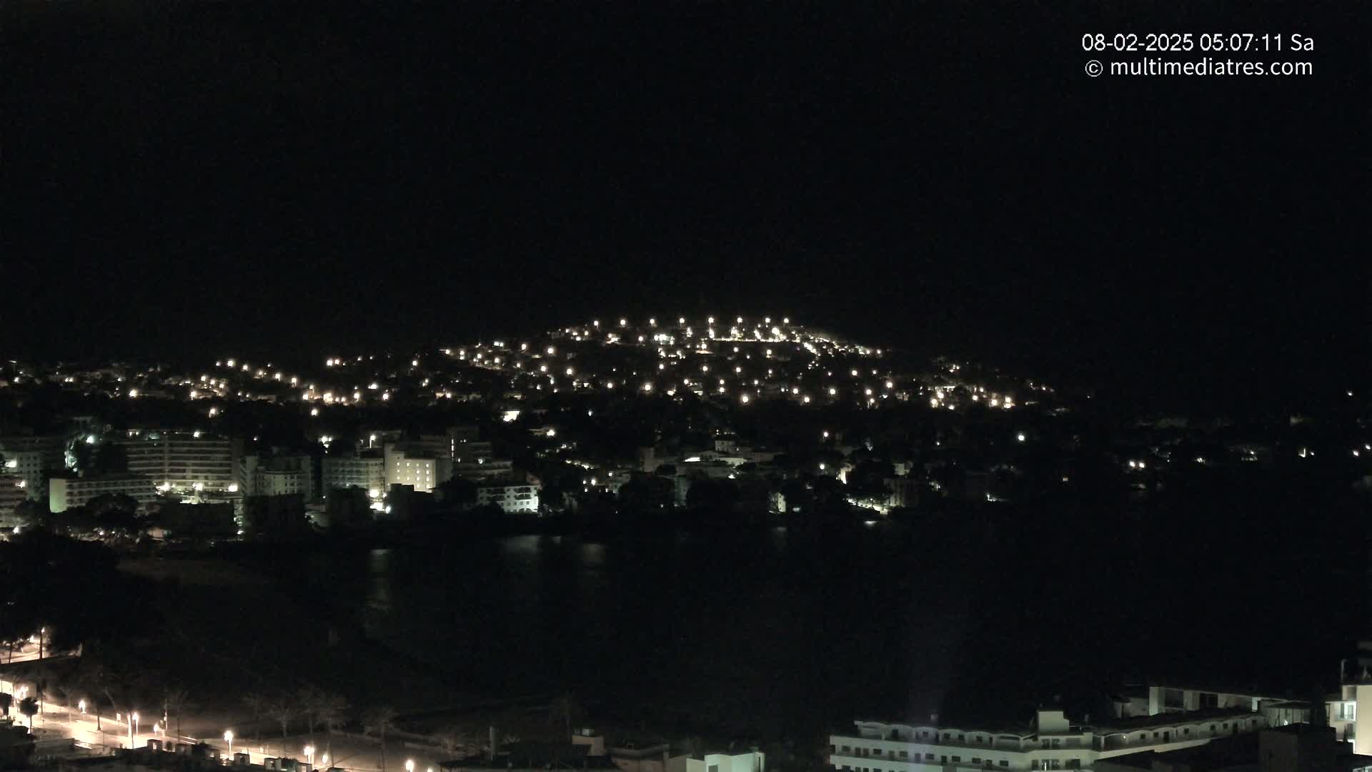 A nighttime view of a coastal town illuminated by scattered lights, with a dark body of water in the foreground.