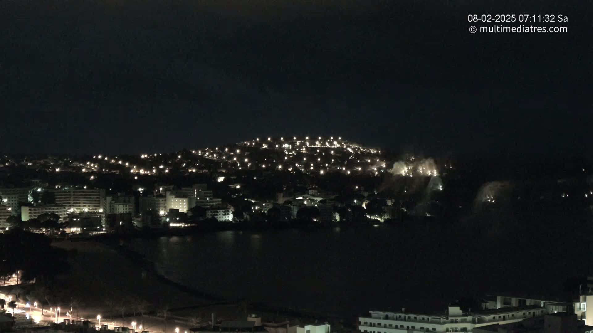 A nighttime view of a city on a hill next to a dark body of water, with some lights appearing to be fireworks or flares in the distance.