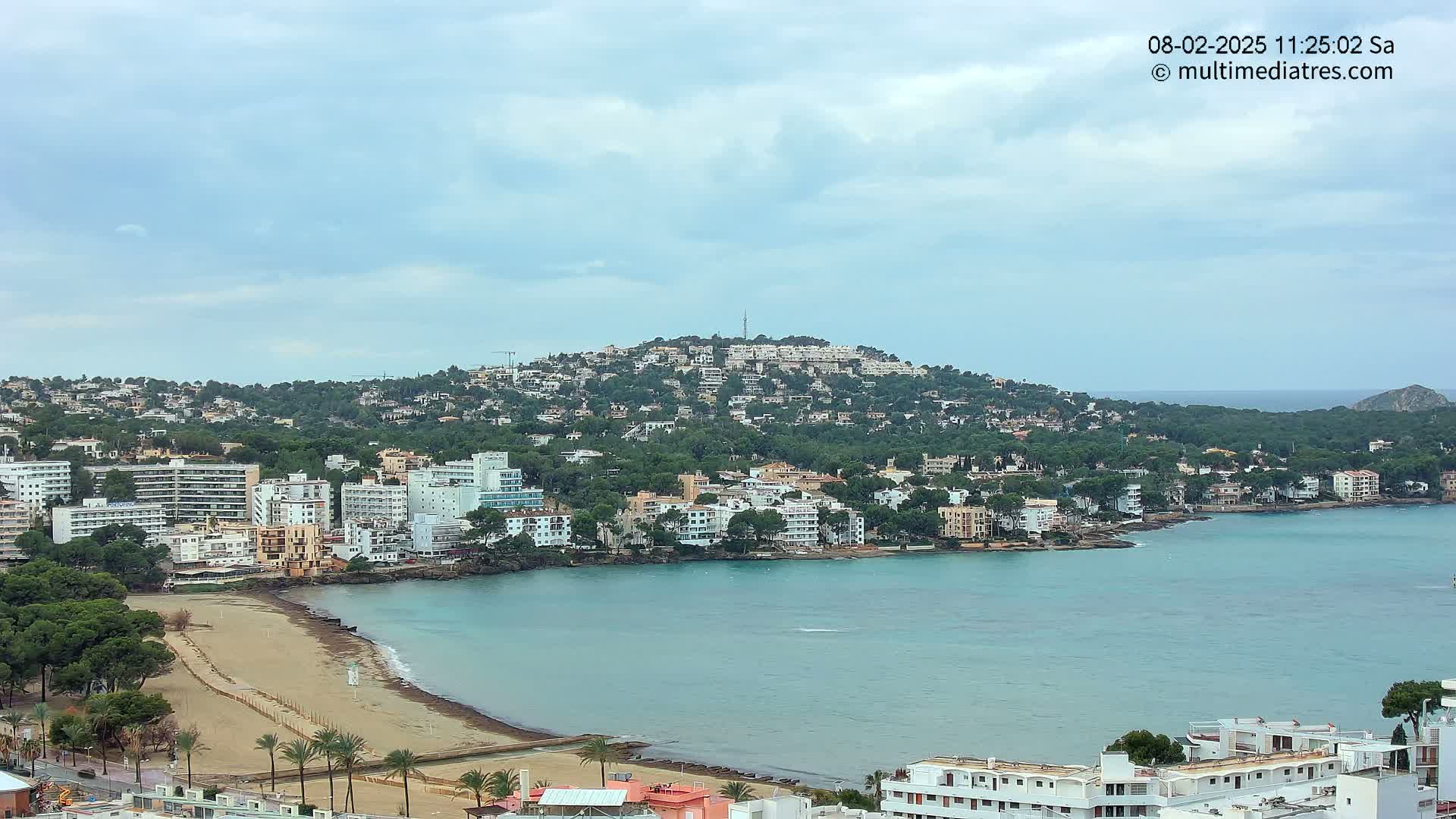 A coastal town with sandy beach and buildings nestled on a hillside under a partly cloudy sky.