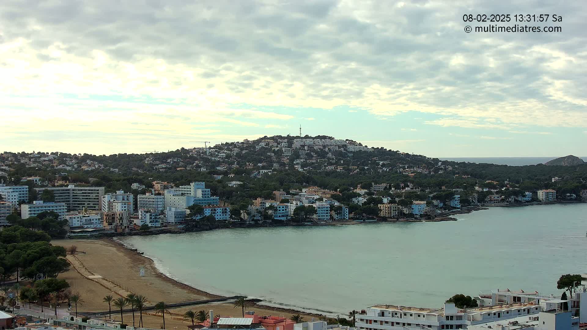 A coastal town with buildings on a hill and along a calm, light-green bay under a partly cloudy sky.