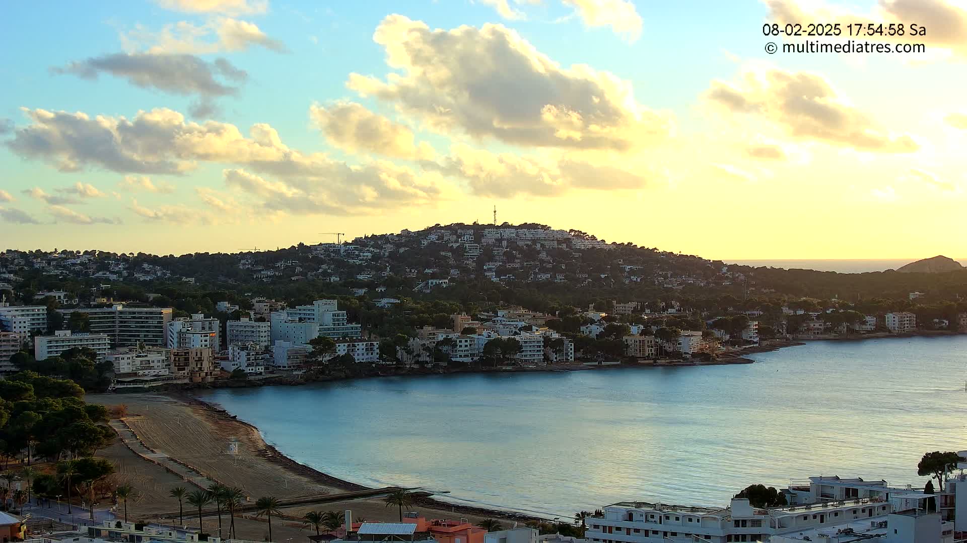 A coastal town nestled against a hill at sunset, under a partly cloudy sky.
