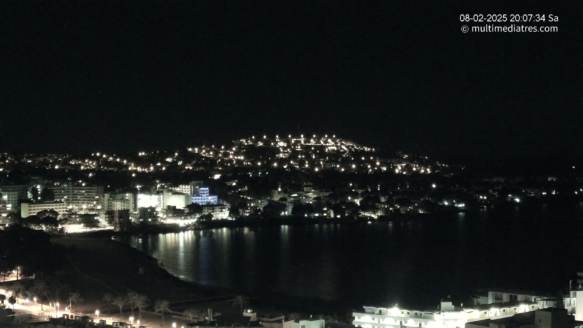 A coastal town is illuminated at night, with its lights reflecting on a dark body of water under a clear, dark sky.