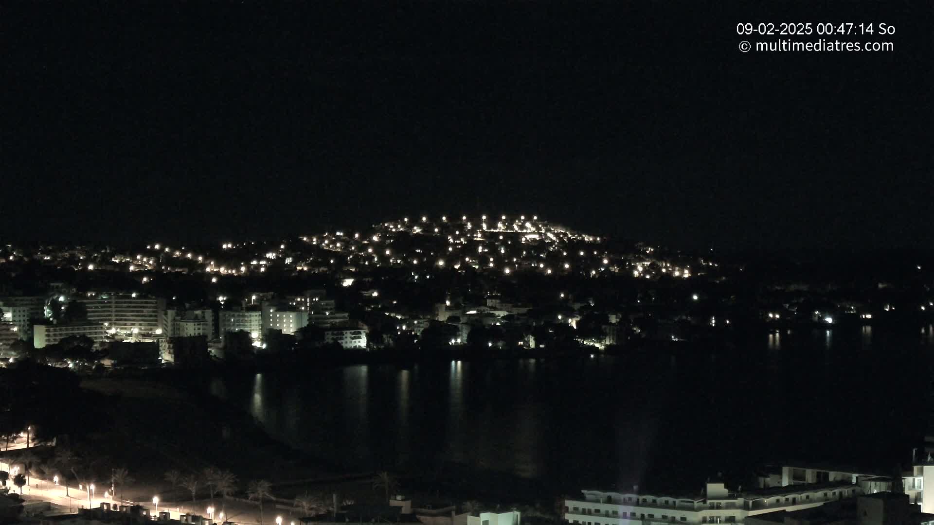 A nighttime view of a city illuminated by lights, situated on a hill overlooking a dark body of water under a clear, dark sky.