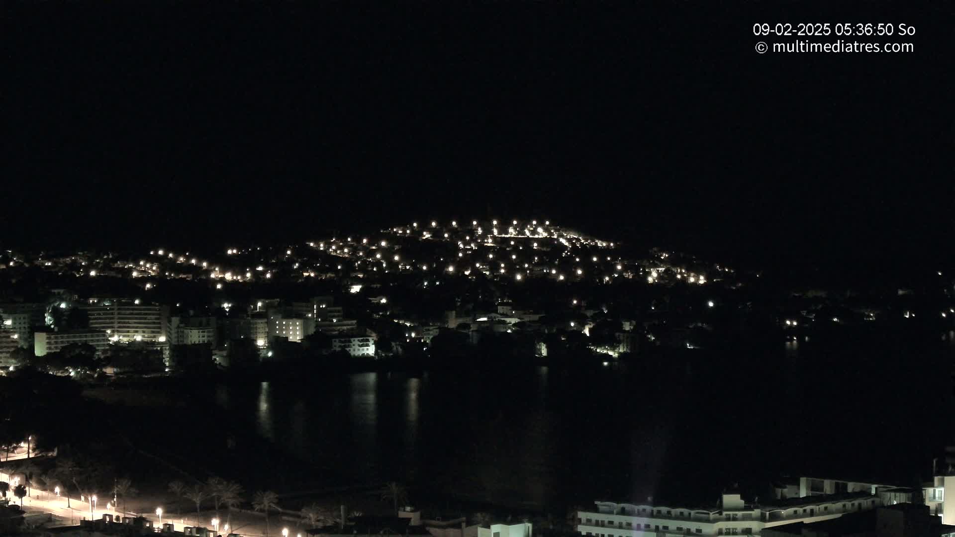 A nighttime view of a coastal city illuminated by many small lights, with dark water in the foreground.