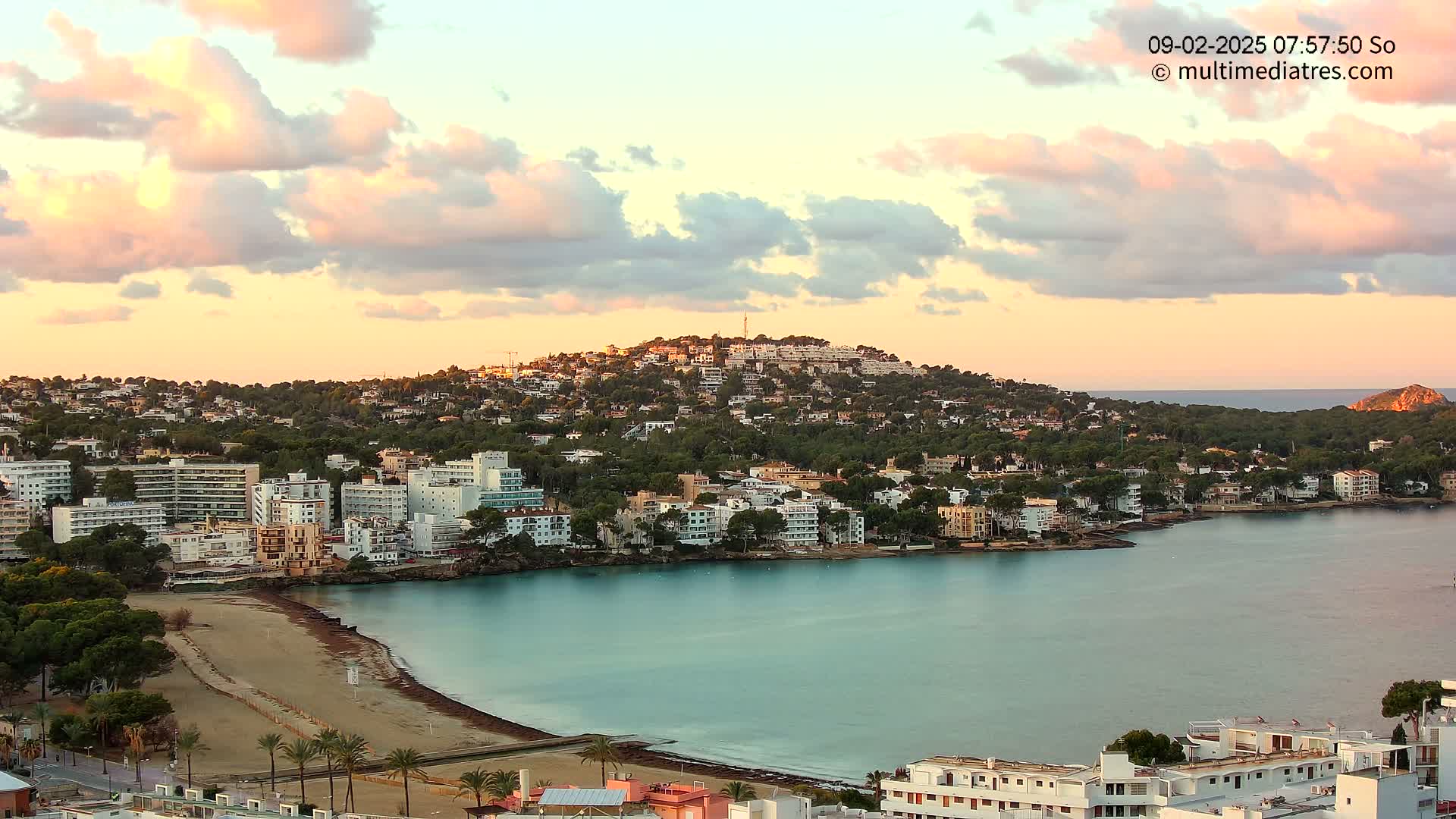 A coastal town nestled against a hill, under a sky filled with pastel-colored clouds at sunrise, features a calm bay with a sandy beach.