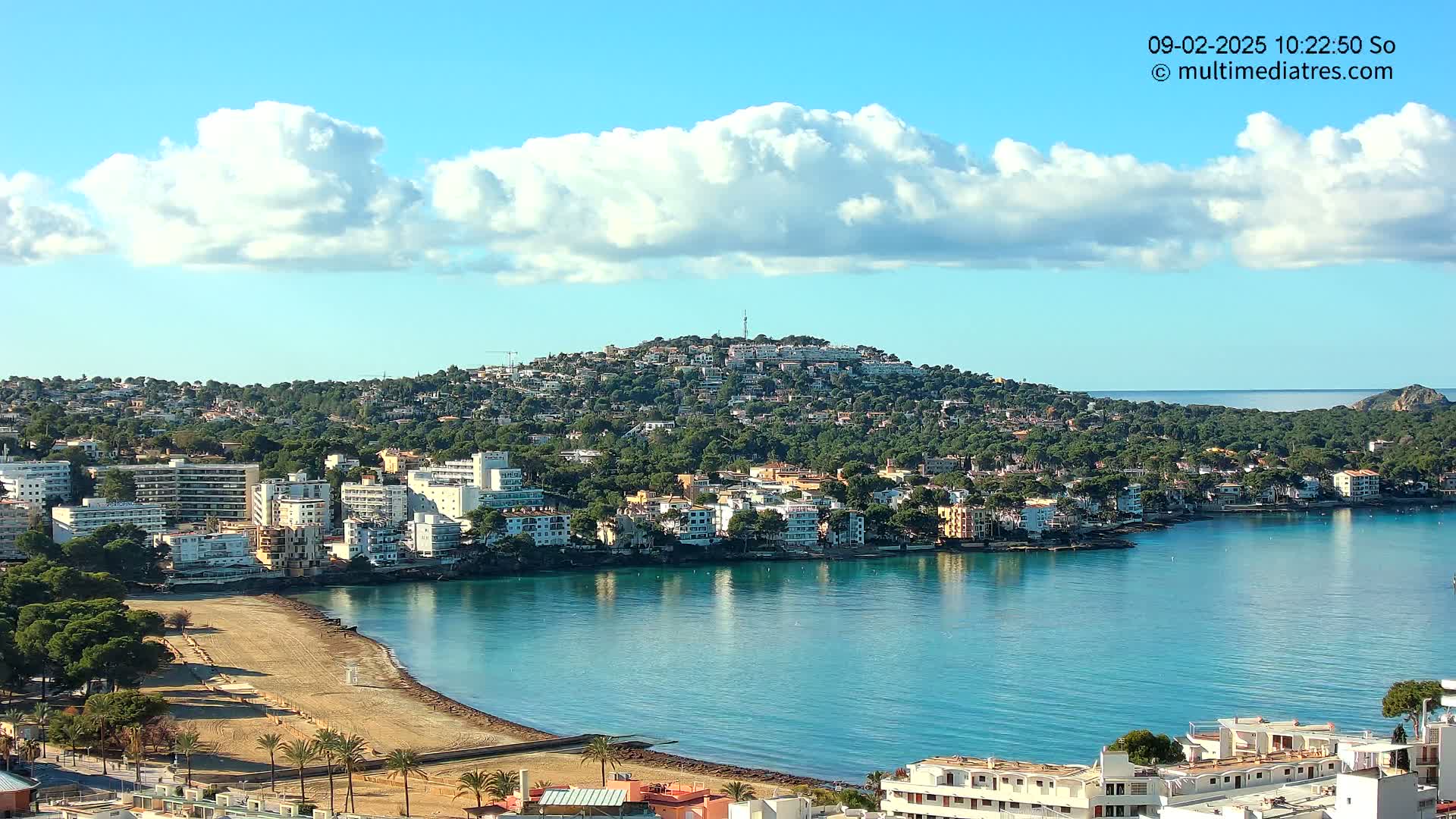 A coastal town with sandy beach and buildings nestled on a hillside under a partly cloudy blue sky.