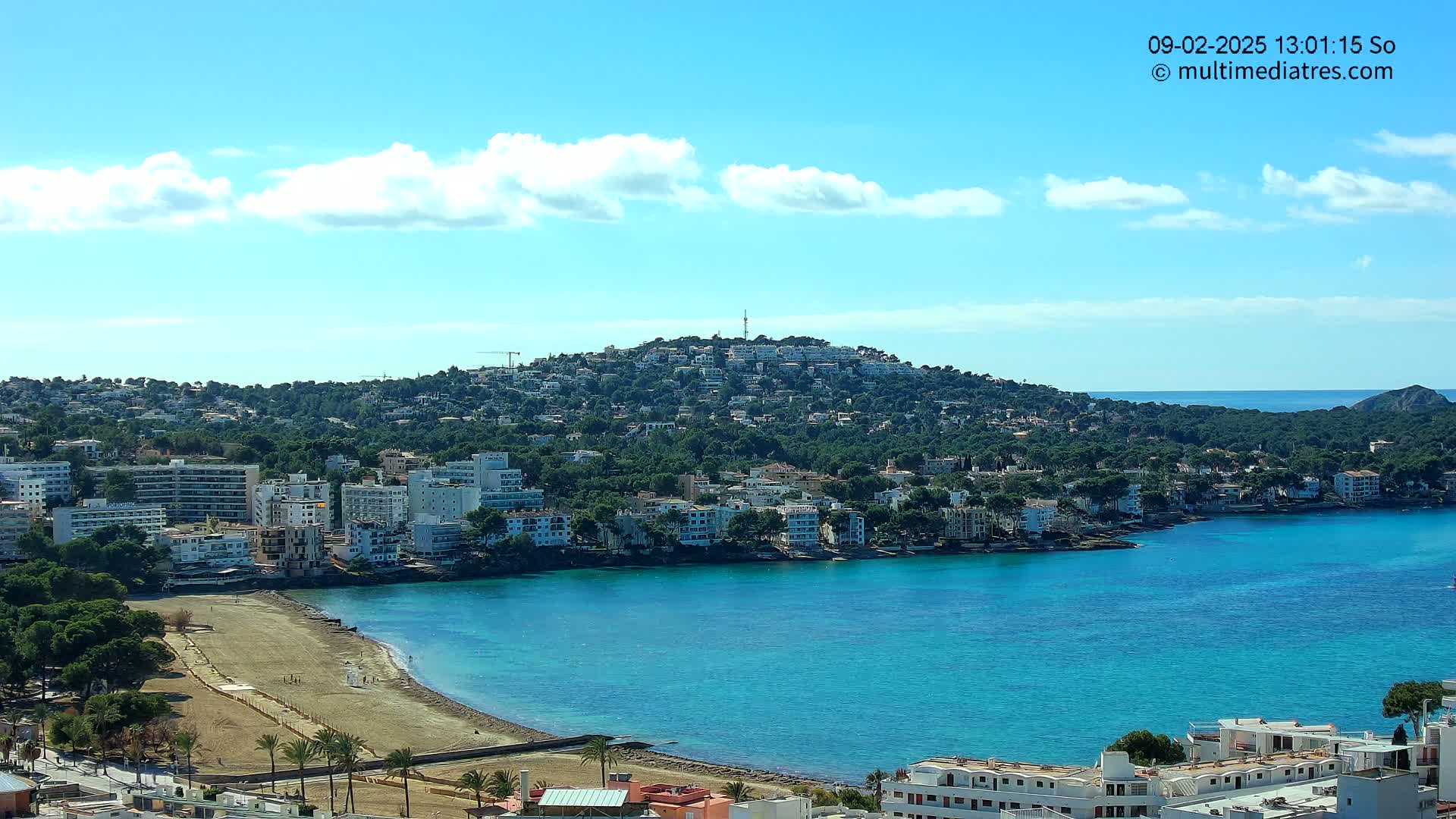 A coastal town nestled on a hill overlooking a calm, turquoise bay under a mostly sunny sky with a few scattered clouds.