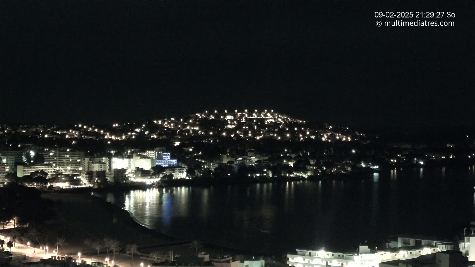 A nighttime view of a coastal town illuminated by many lights, reflected in a dark body of water under a clear, dark sky.