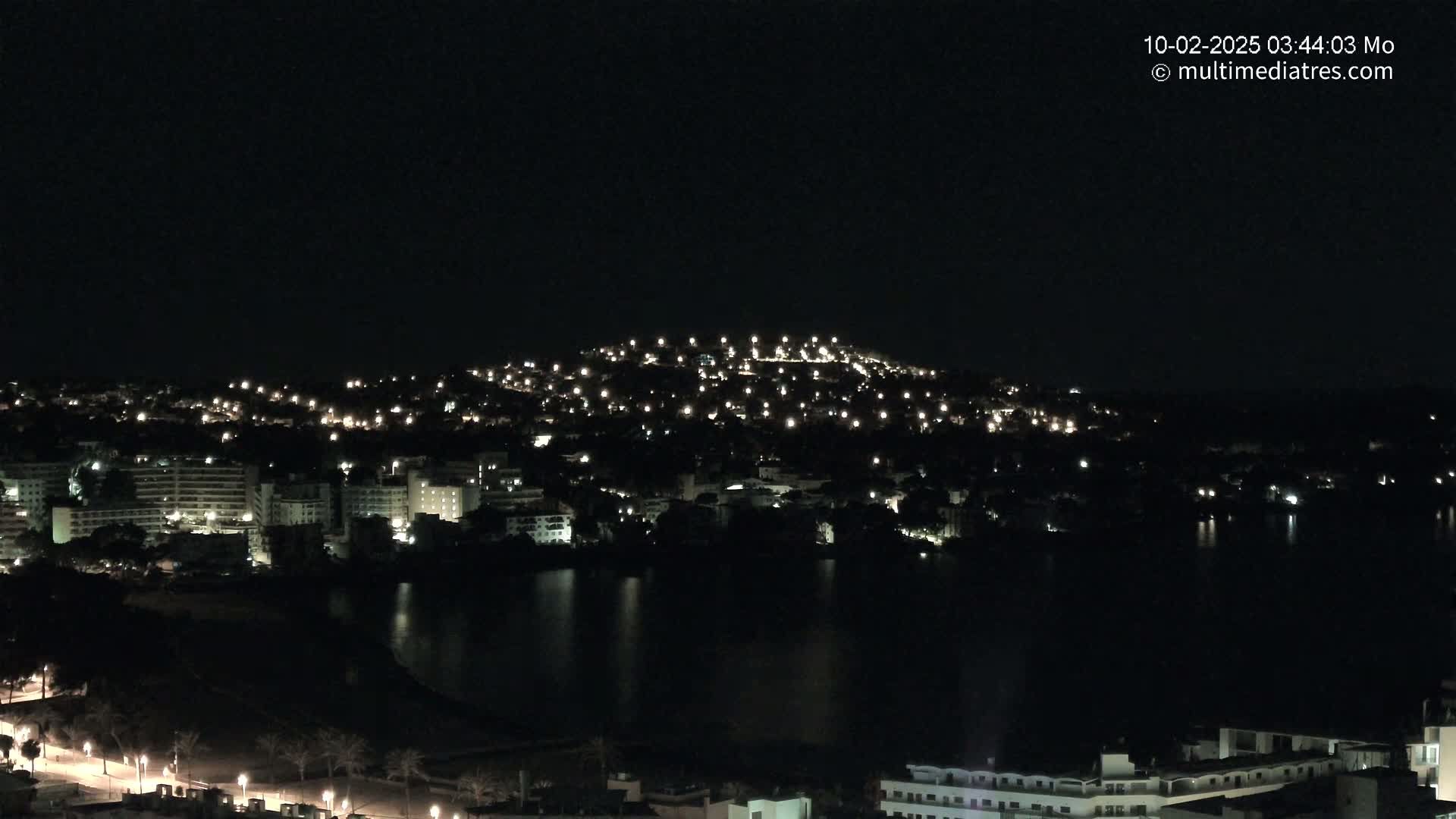 A nighttime view of a city illuminated by lights, nestled beside a dark body of water under a clear, dark sky.
