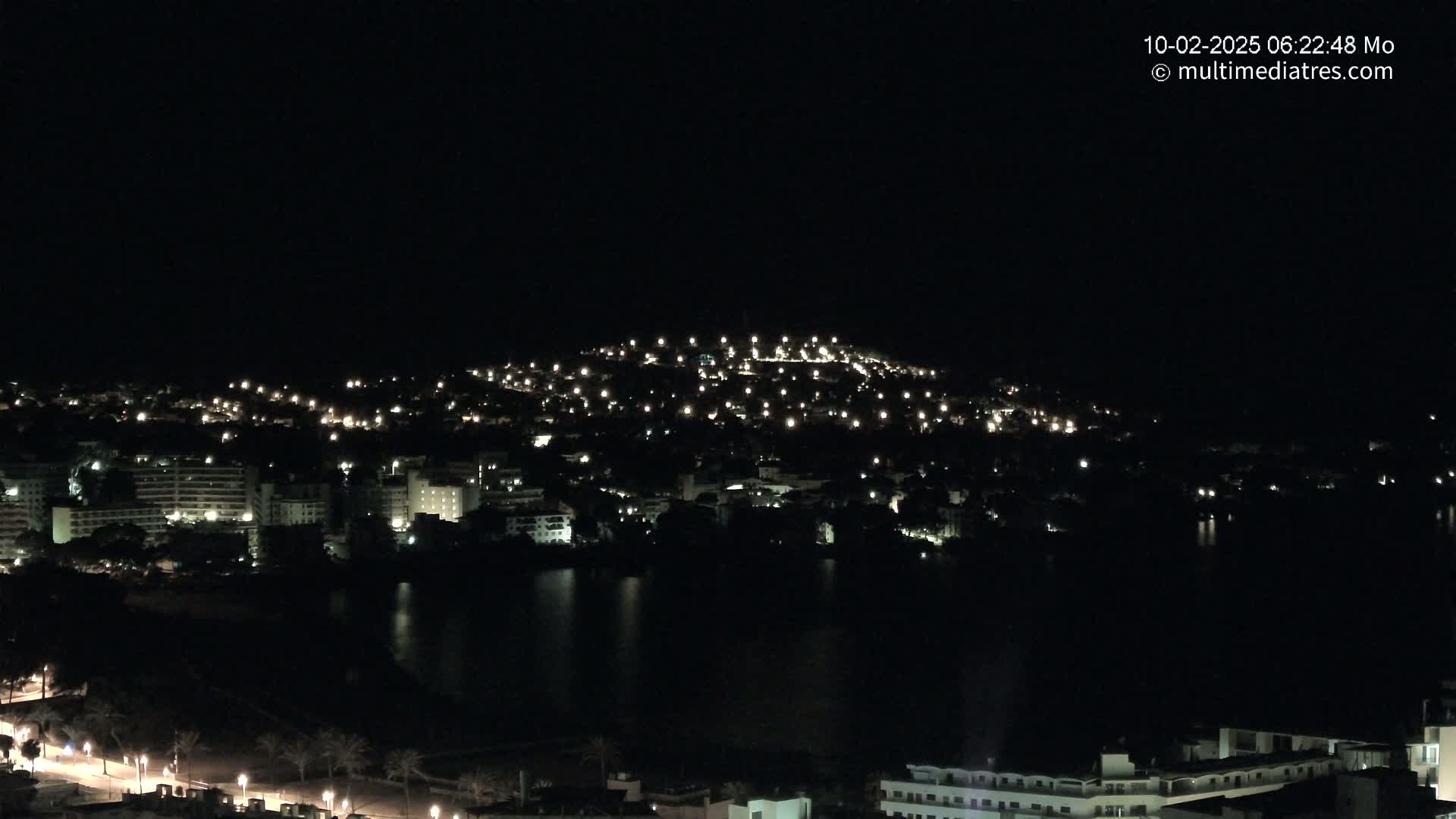 A nighttime view of a city illuminated by scattered lights, situated beside a dark body of water under a clear, dark sky.