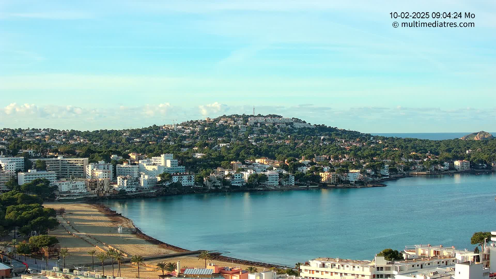 A coastal town nestled against a verdant hill, with a sandy beach curving into a calm, blue bay under a mostly clear sky.