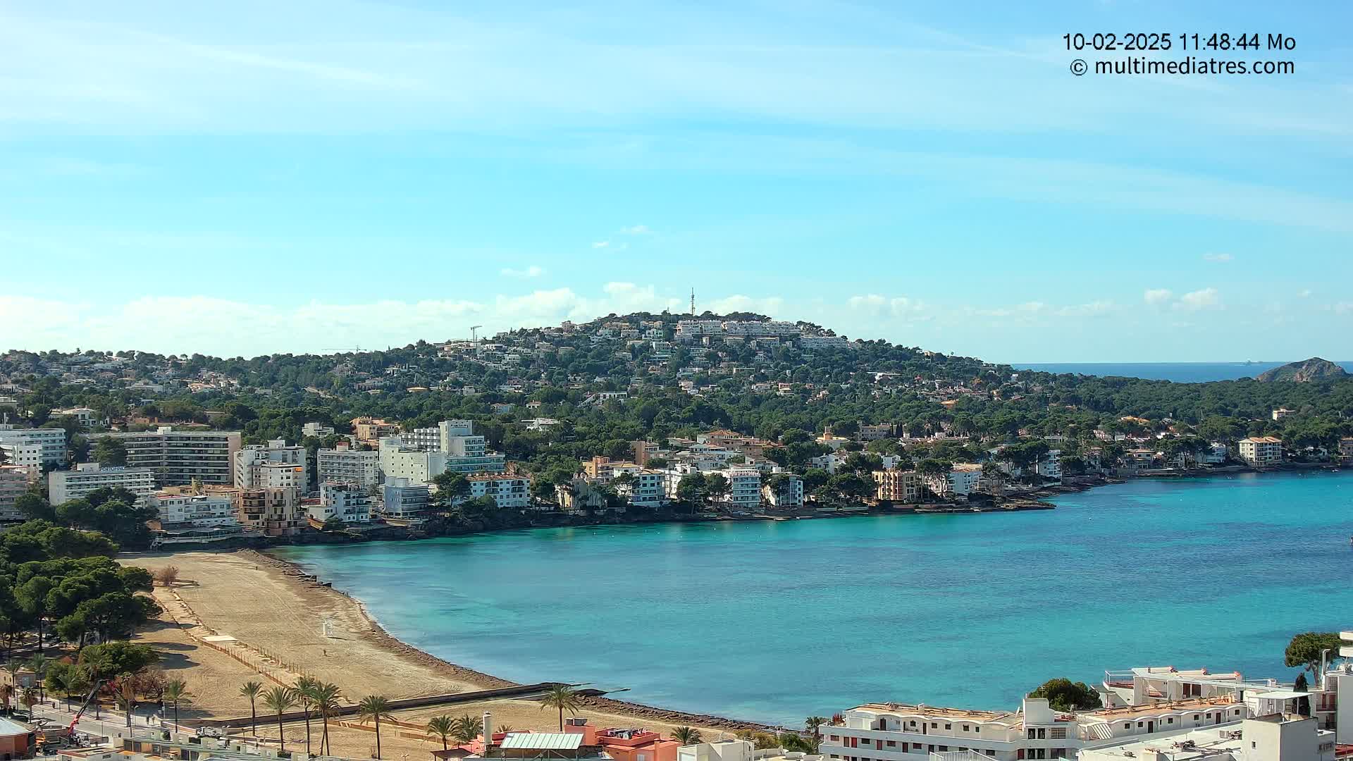 A coastal town nestled against a hill overlooking a calm, turquoise bay under a mostly clear blue sky.