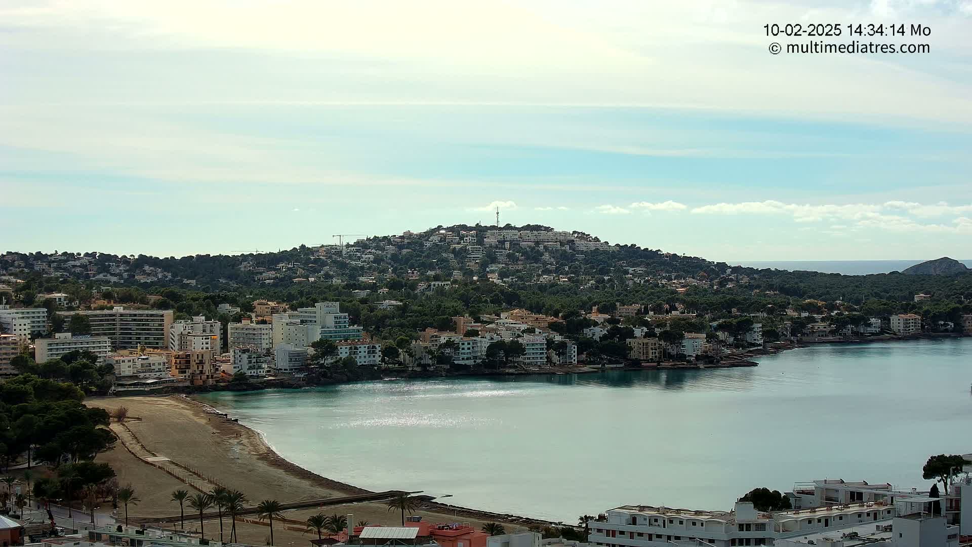 A coastal town with buildings and trees sprawls across a hill overlooking a calm, turquoise bay under a partly cloudy sky.