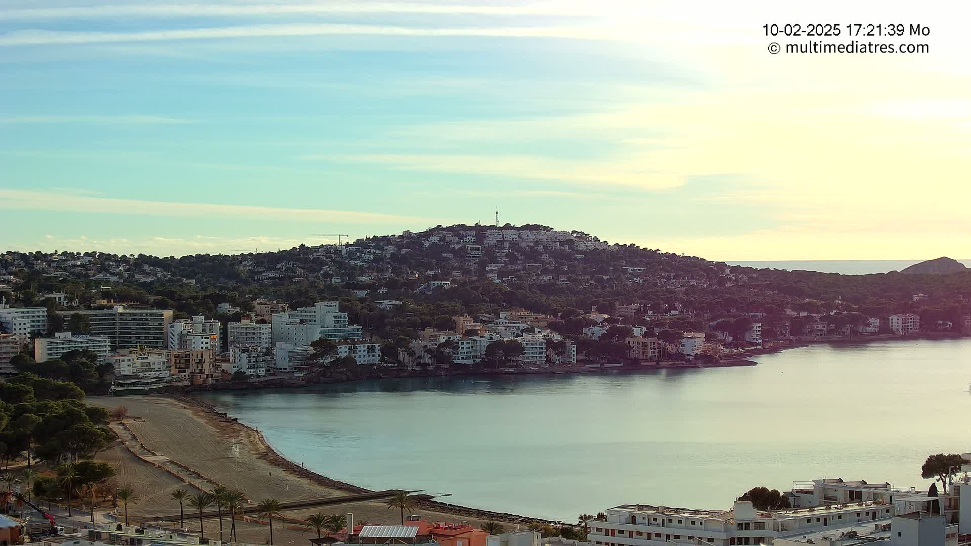 A coastal town nestled on a hill overlooking a calm bay under a clear, light blue sky.